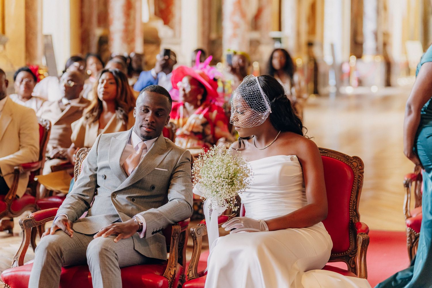 Wedding guests in formal attire watch a celebration in an elegant venue with ornate decor and warm lighting.