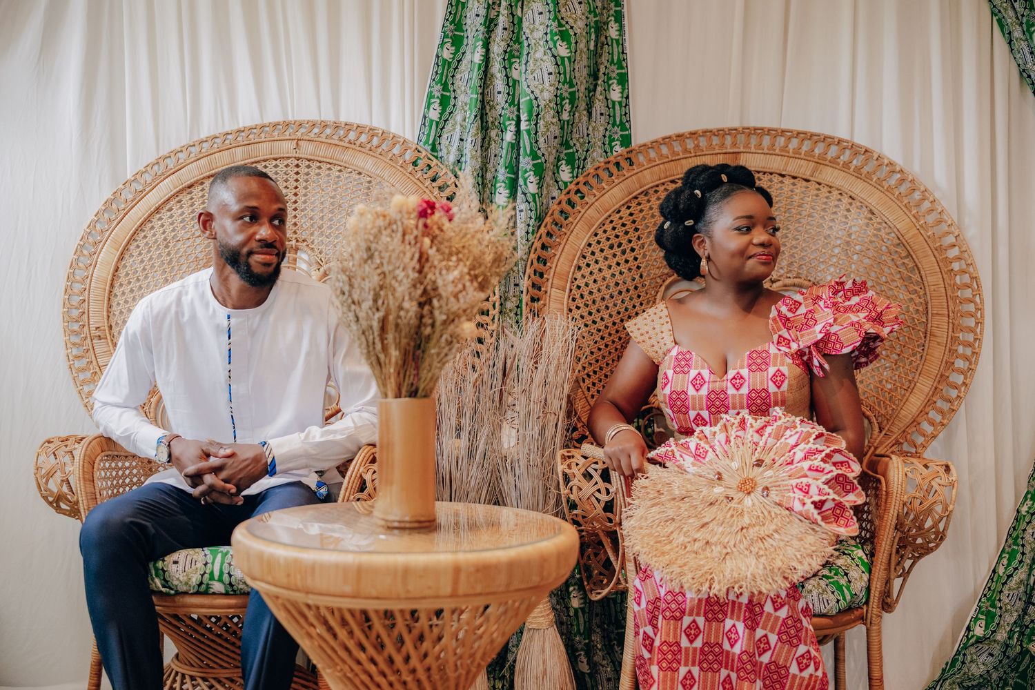 Wedding reception setup with woven peacock chairs and green drapery decorated with pink floral accents.
