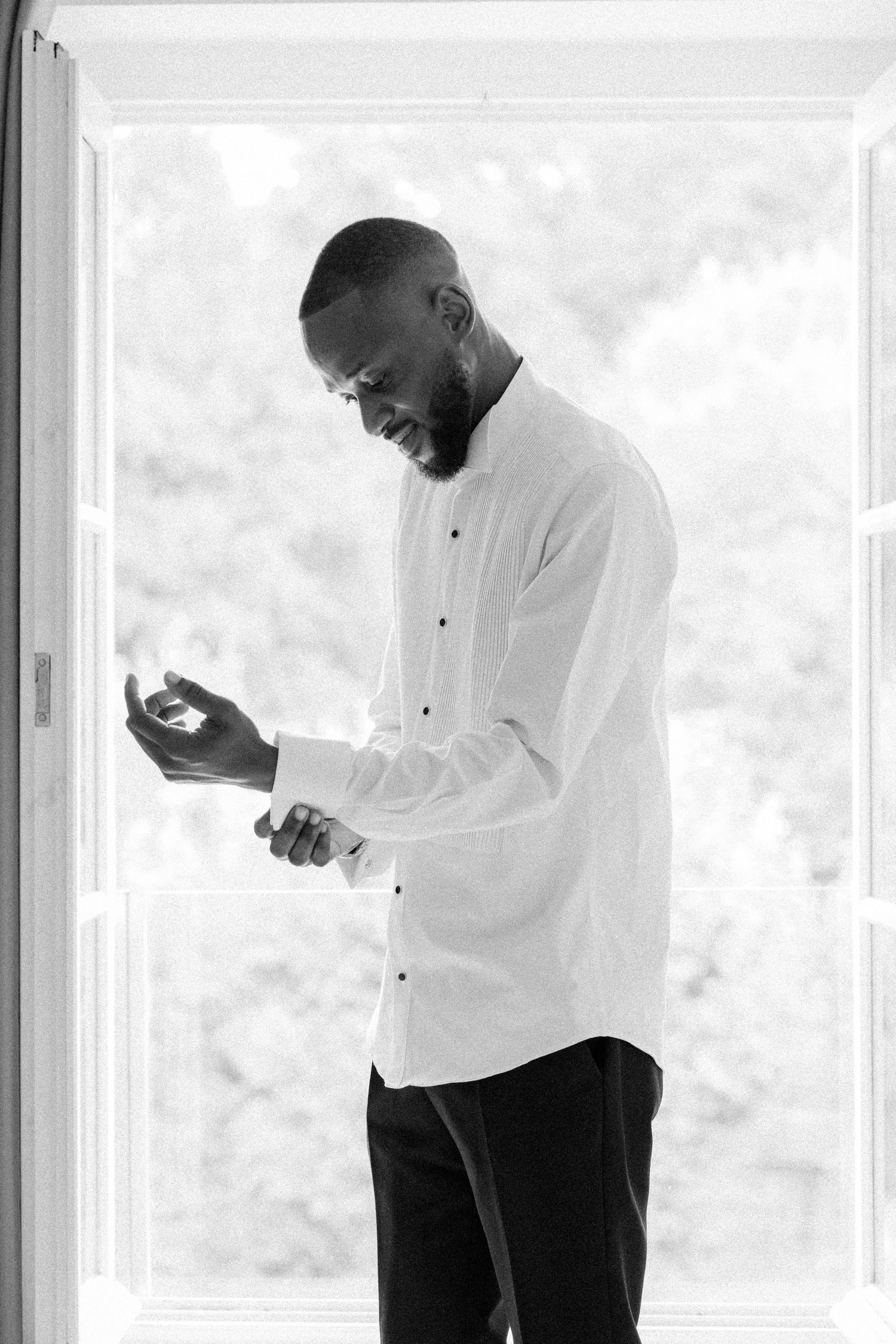 A person in a white dress shirt and black pants stands near a window in a bright room.
