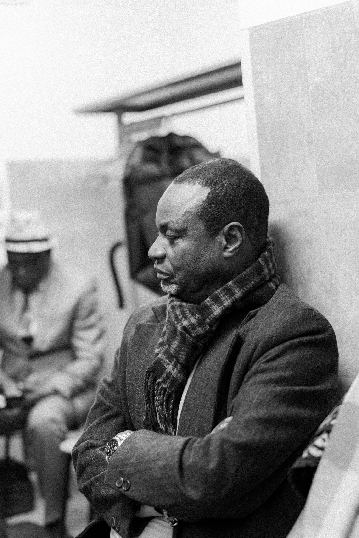 A black and white image shows a person wearing a winter coat and scarf sitting against a wall.