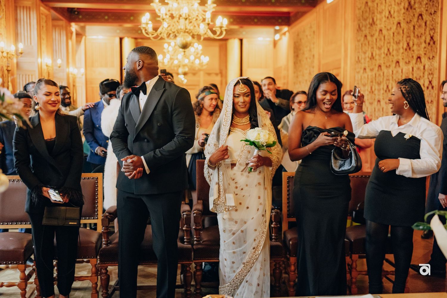 A group of formally dressed guests gathered in an ornate room with a crystal chandelier.