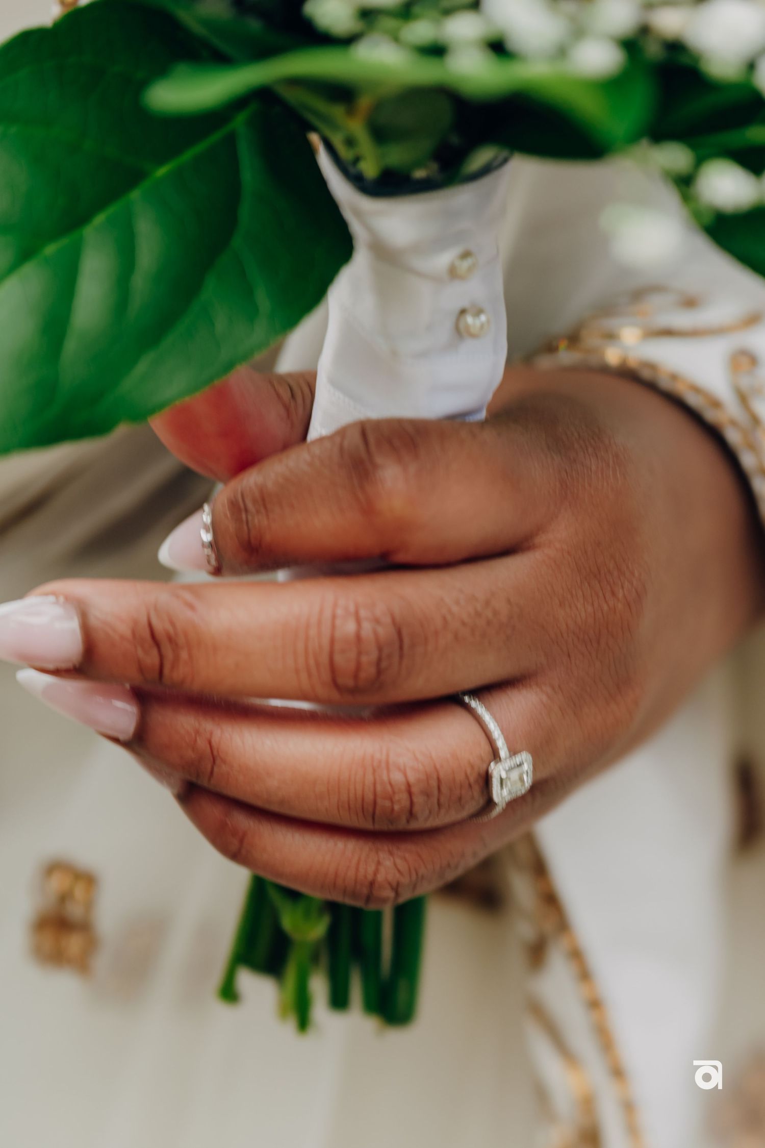 Close-up of diamond engagement ring with green foliage and bouquet in background.
