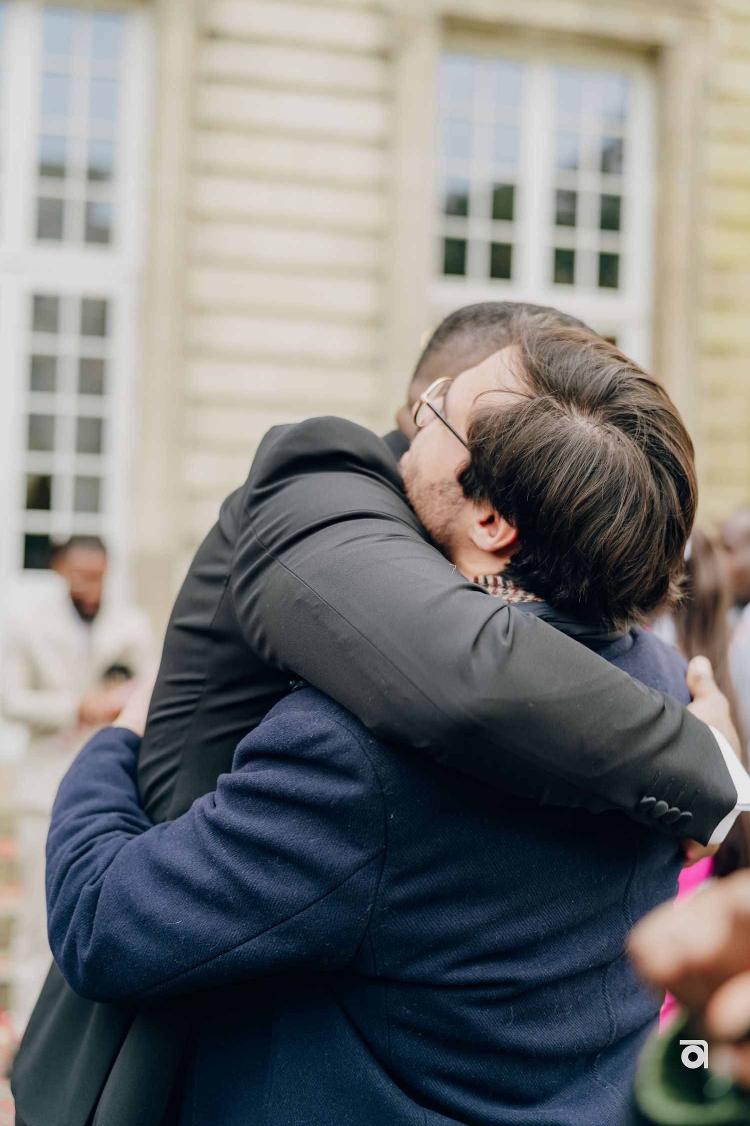 Two people embrace in a warm hug outside a grand building with classical architecture.