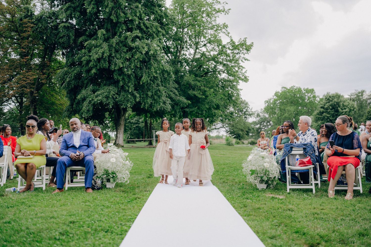 Wedding guests sit in white chairs on a grassy hillside during an outdoor summer ceremony.