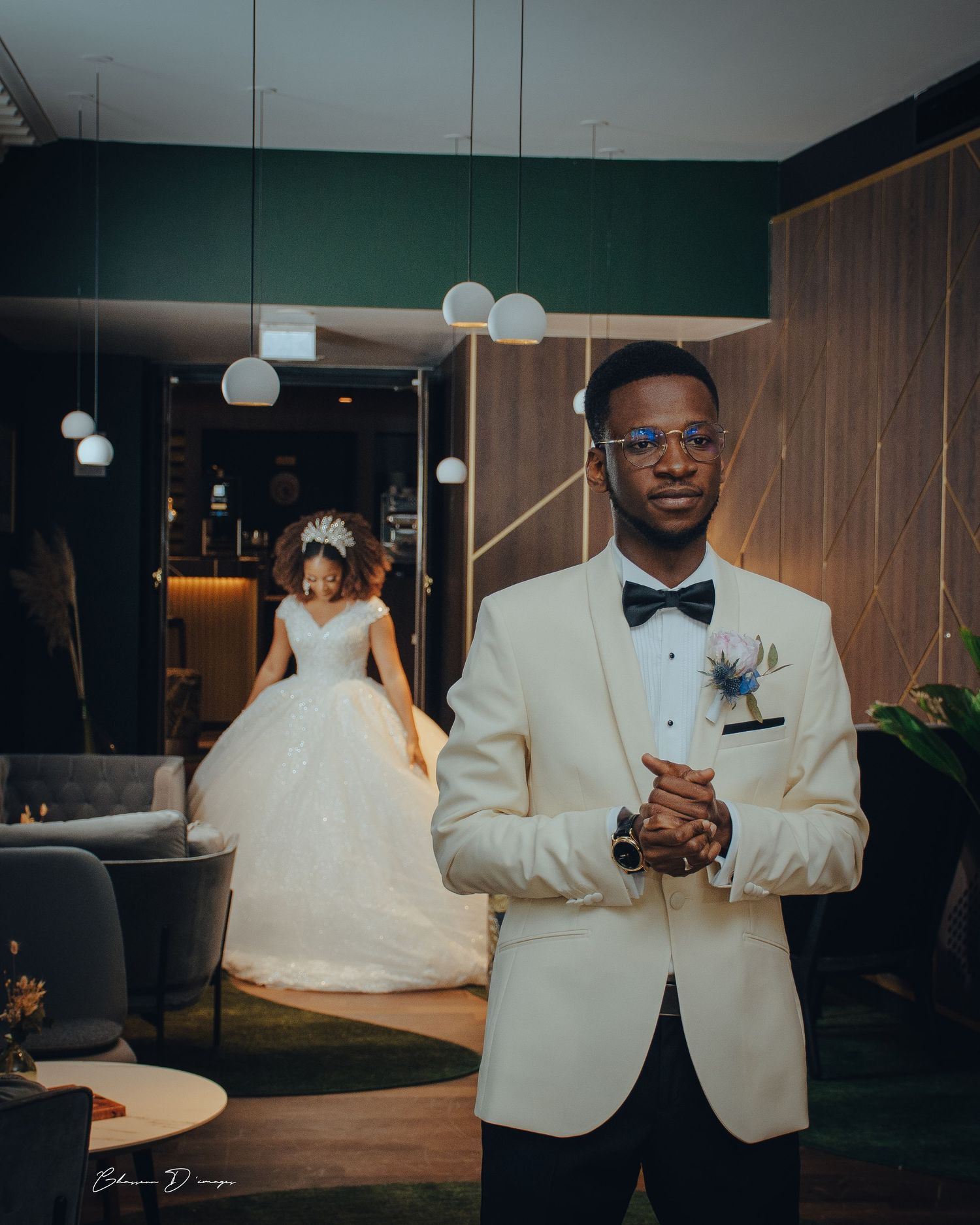 A groom in white tuxedo stands in modern venue with pendant lights.