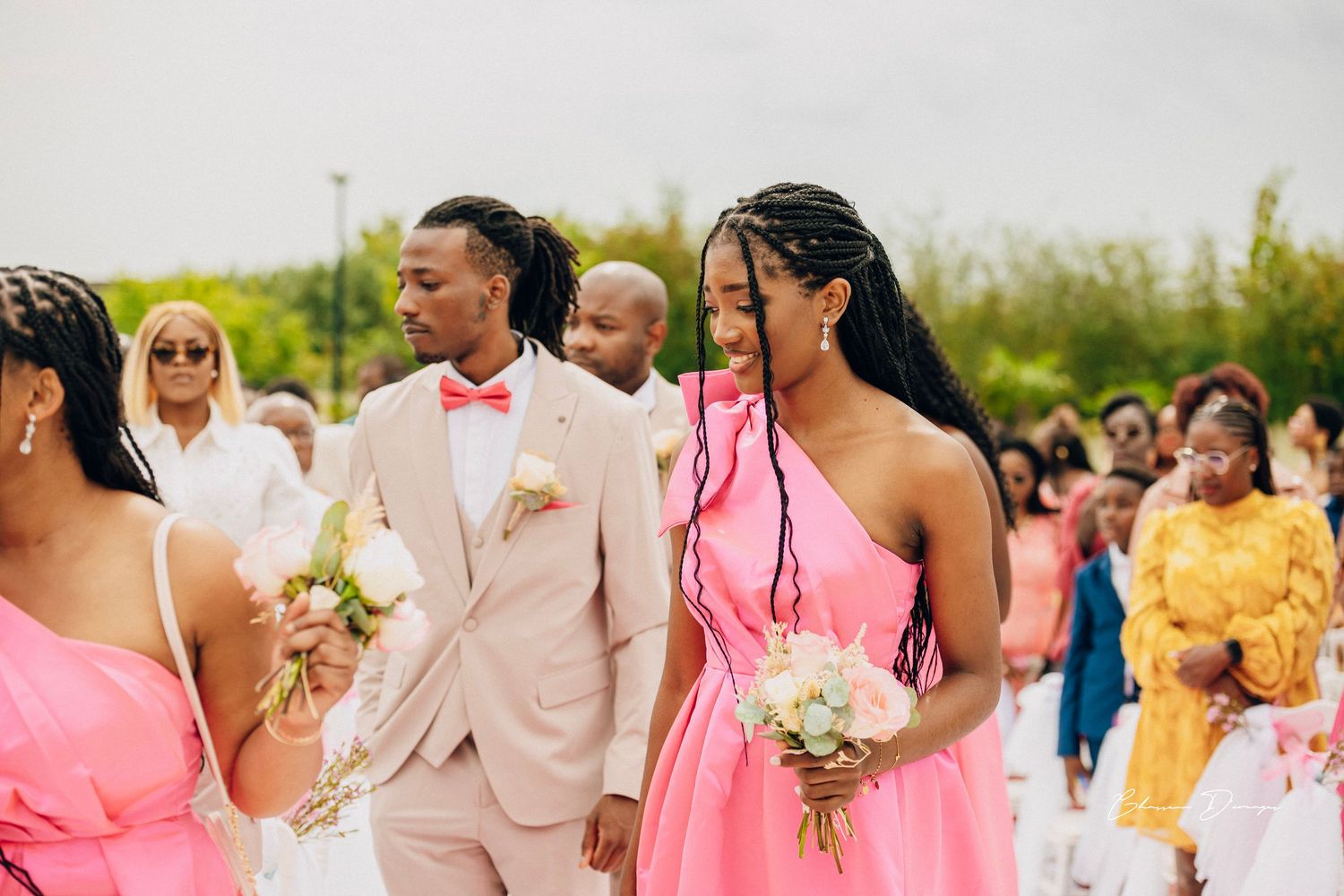 Wedding party members in pink dresses and beige suits walk together during an outdoor ceremony.