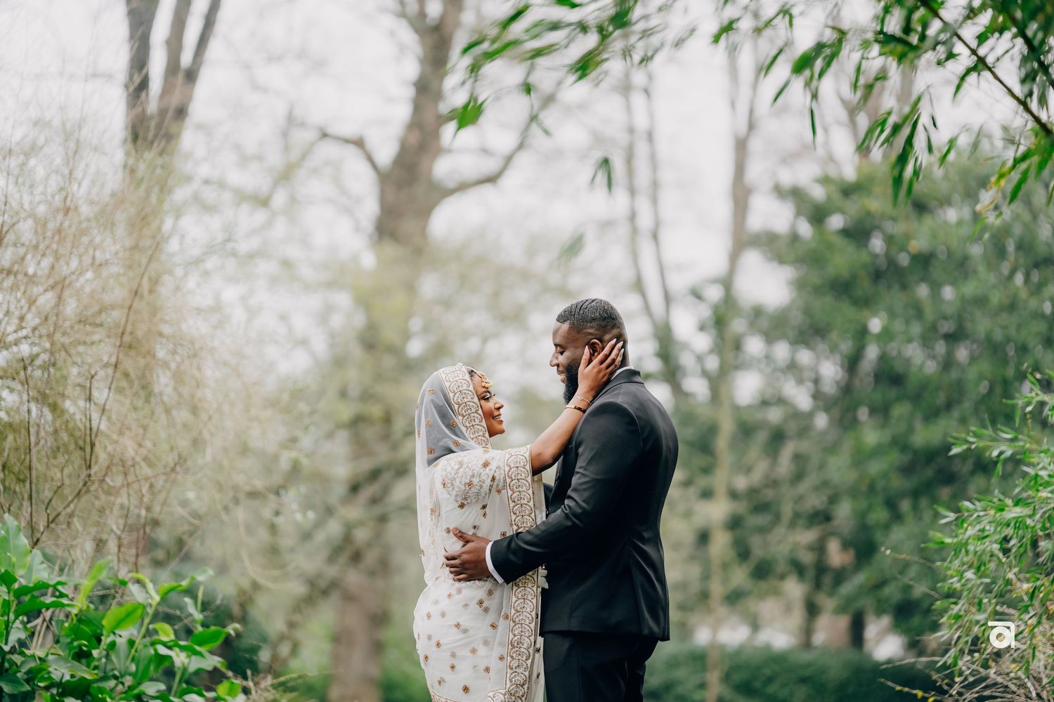 A romantic outdoor wedding photo of a bride in a white lace dress and groom in a black suit among trees and greenery.