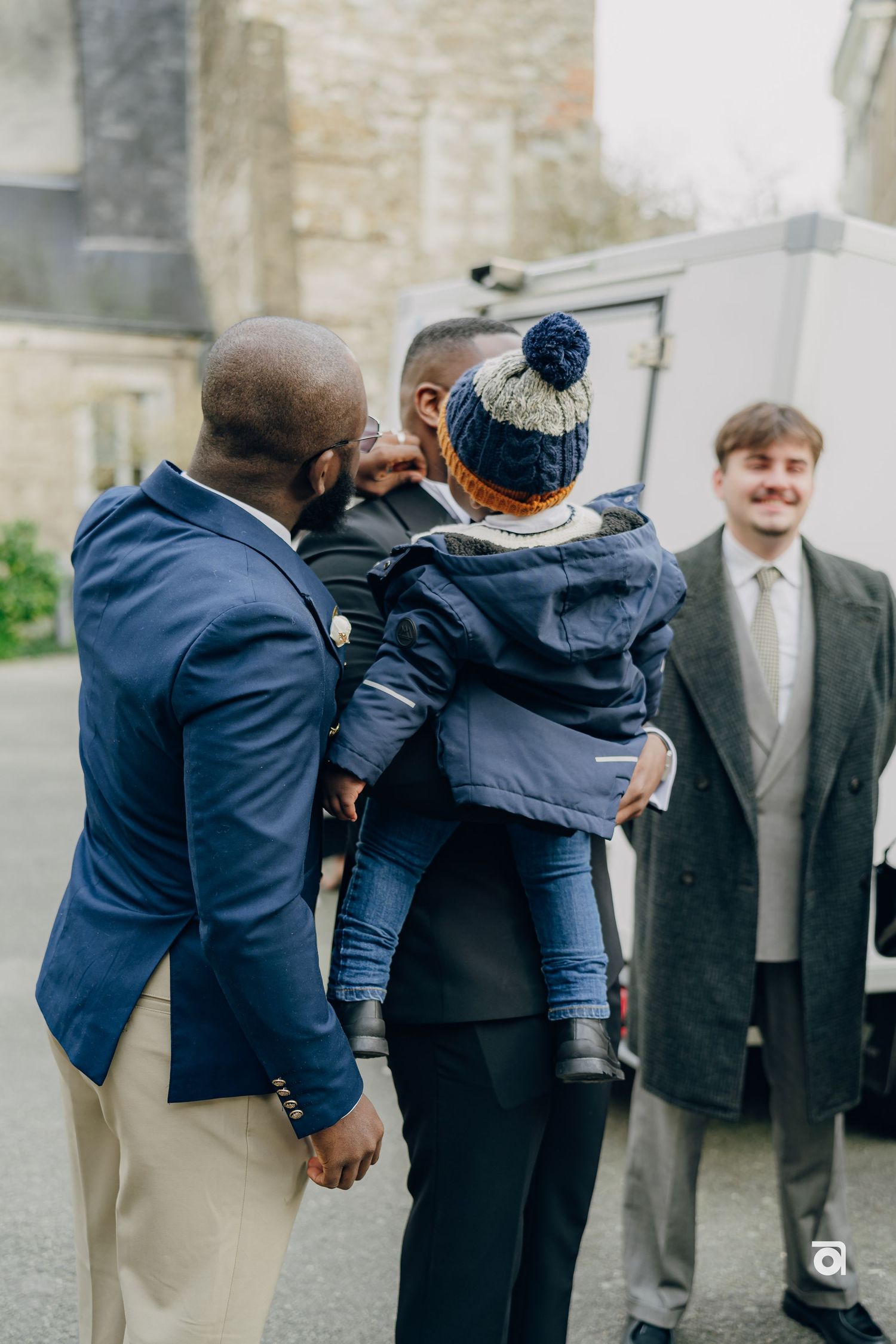 A small child in a winter hat is held up while interacting with others outside a building.