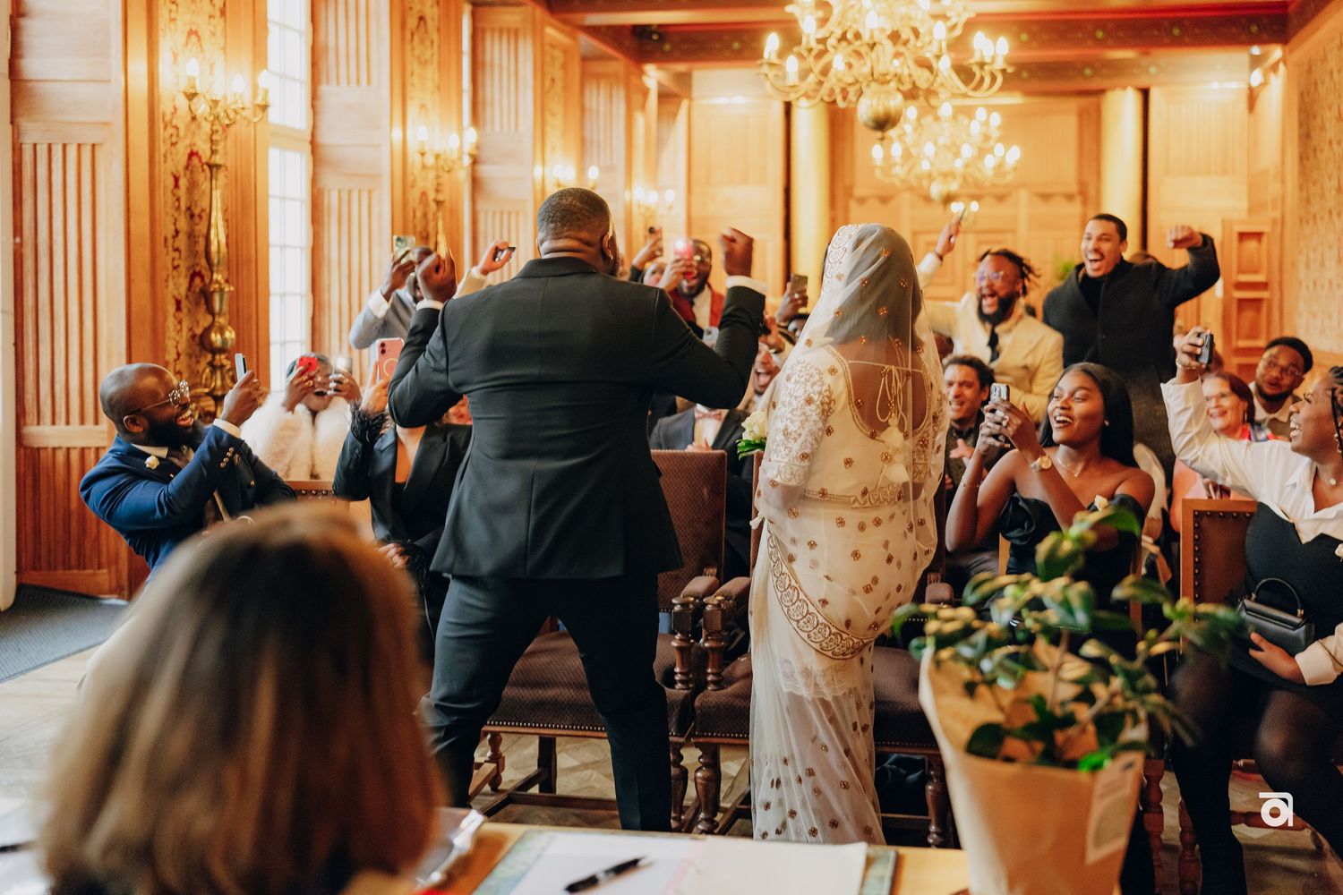 Wedding celebration in an ornate venue with chandelier as guests cheer and applaud.