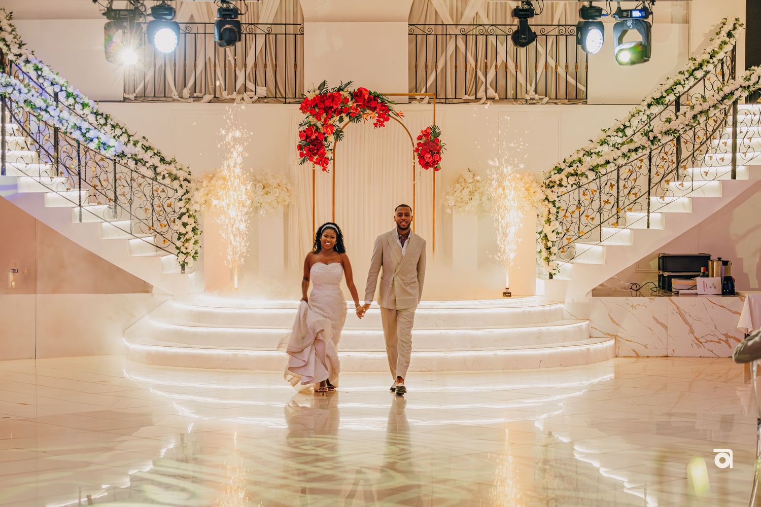 Two people hold hands while descending an elegant white staircase adorned with holiday lights and red poinsettias.