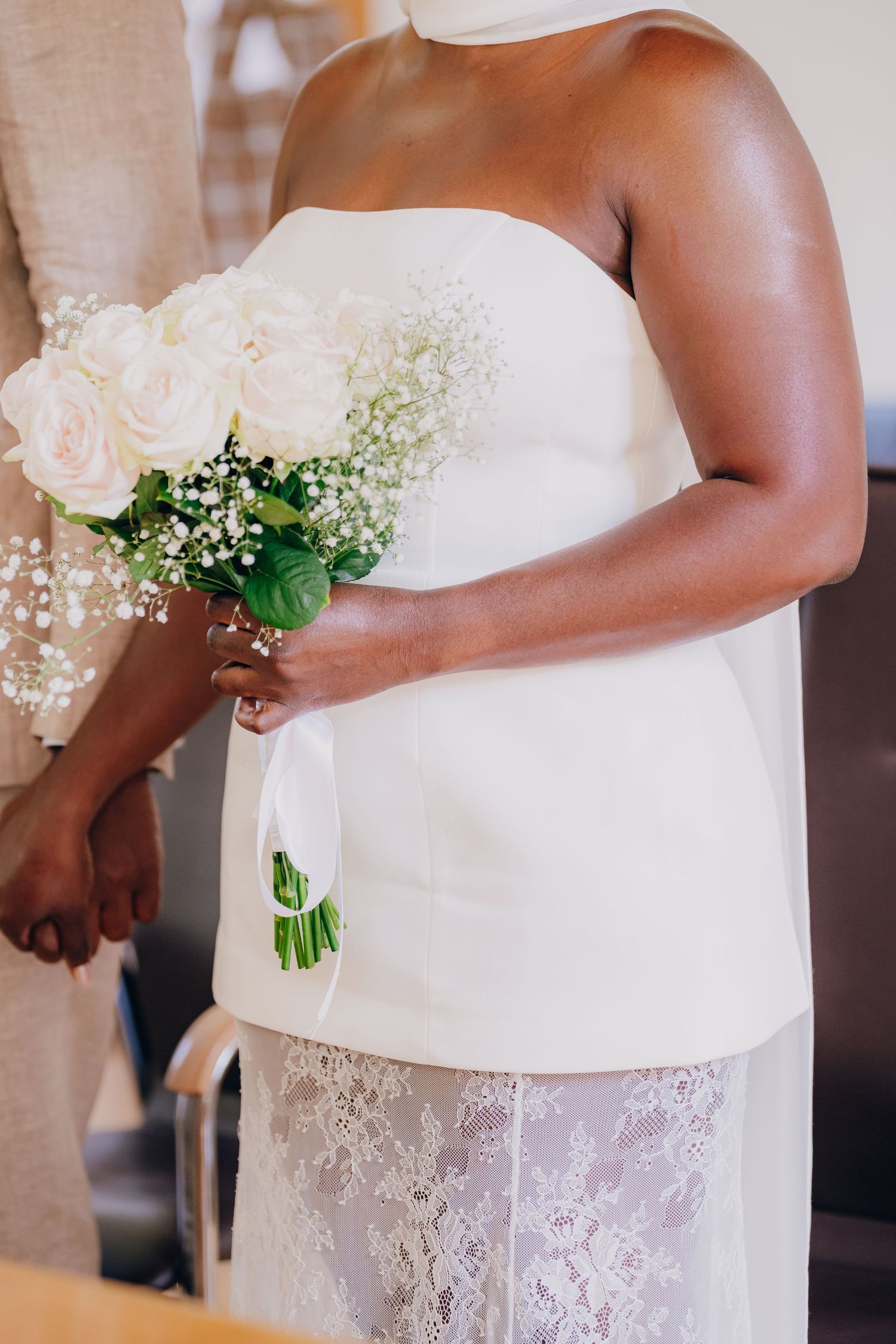 A bride in a white lace dress holds a delicate bouquet of pink roses and baby's breath flowers.