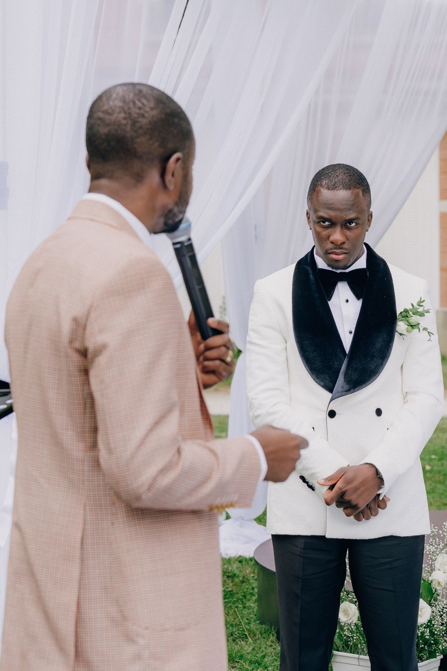 Wedding ceremony participants in formal attire standing under white draping outdoors.