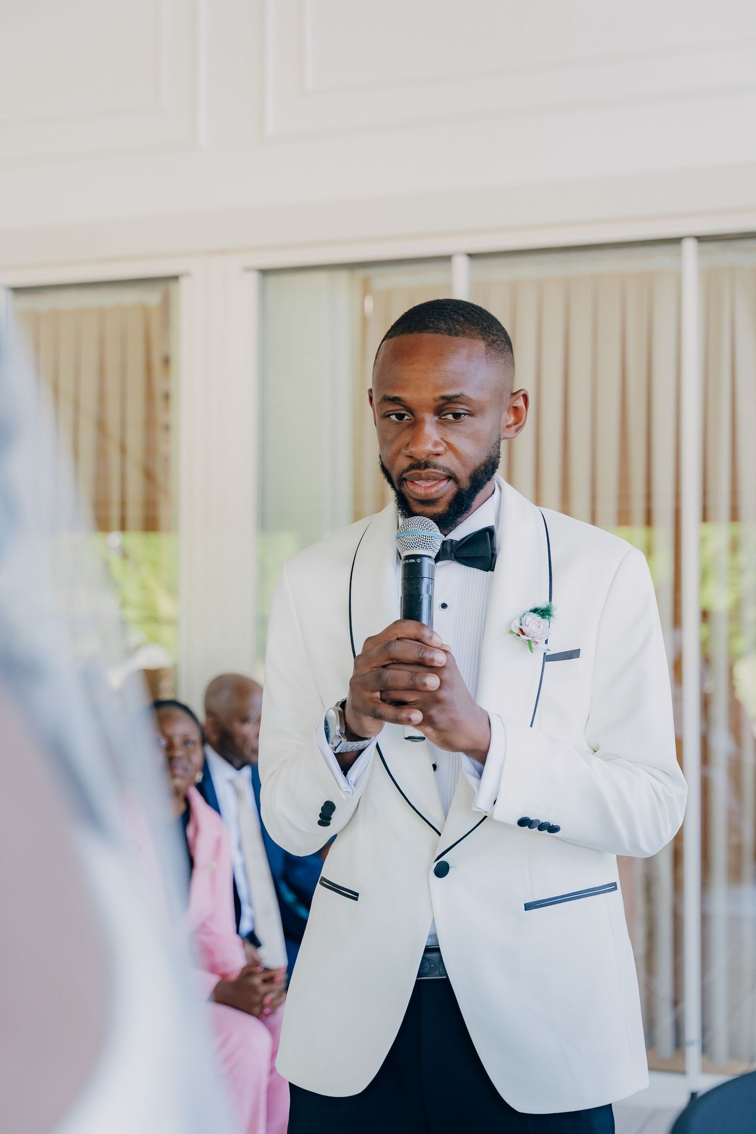 Groom in white tuxedo jacket speaks into microphone during wedding ceremony.