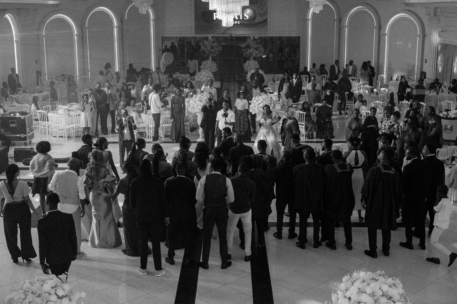 Black and white photo of a crowded wedding reception with guests mingling under elegant arched architecture.