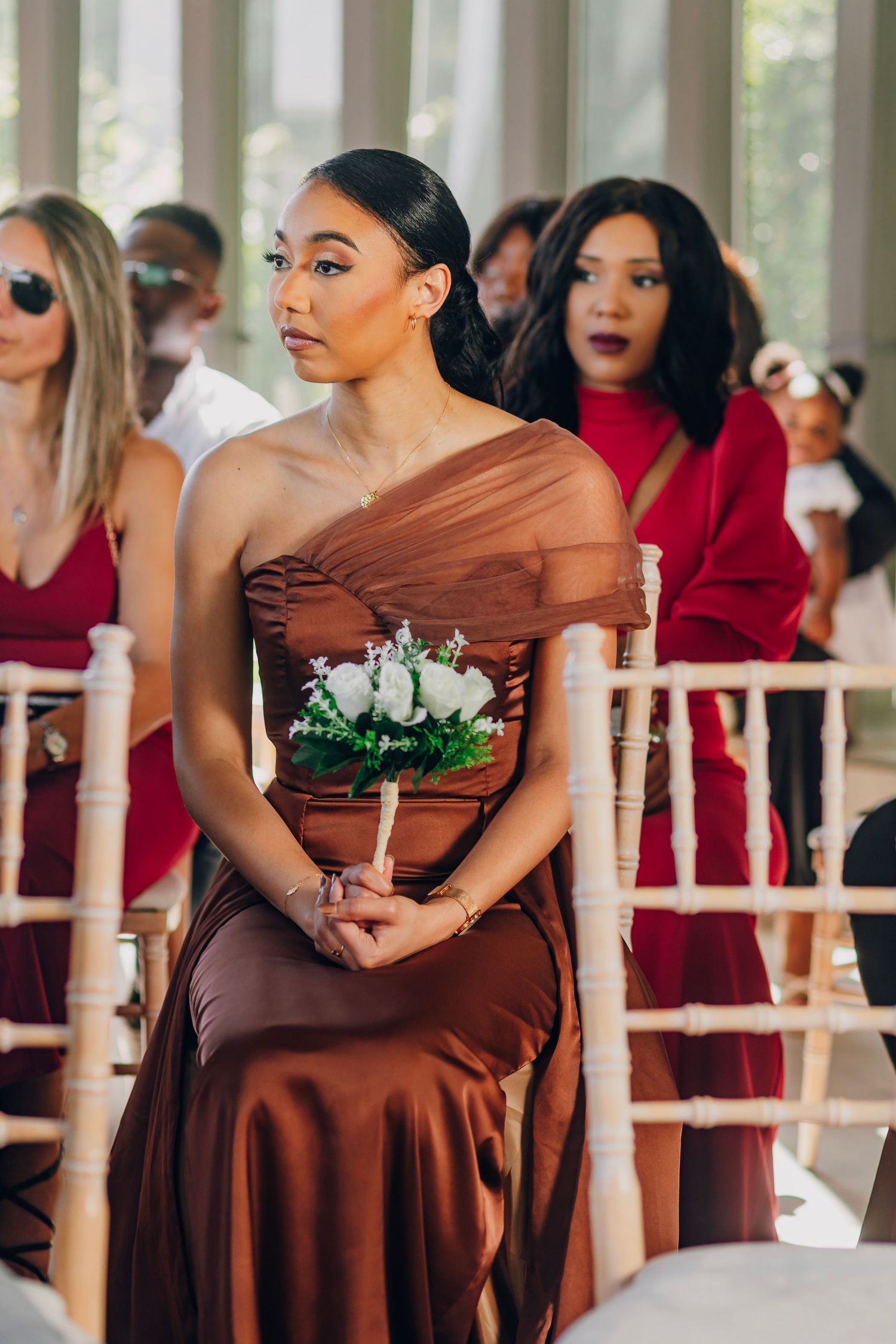 A bridesmaid in a flowing brown dress holds a white flower bouquet at an elegant indoor ceremony.