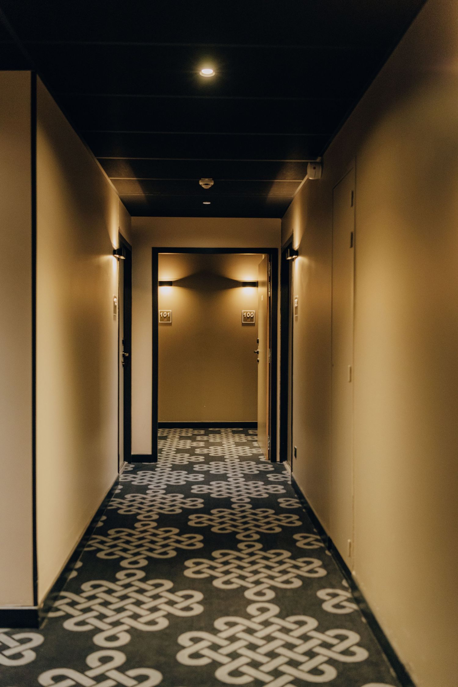A dimly lit hotel hallway with geometric patterned carpet and modern wall sconces casting warm ambient light.