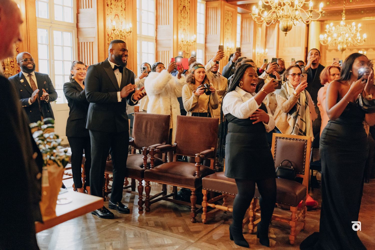 Wedding guests in formal attire celebrate enthusiastically in an elegant room with chandeliers.