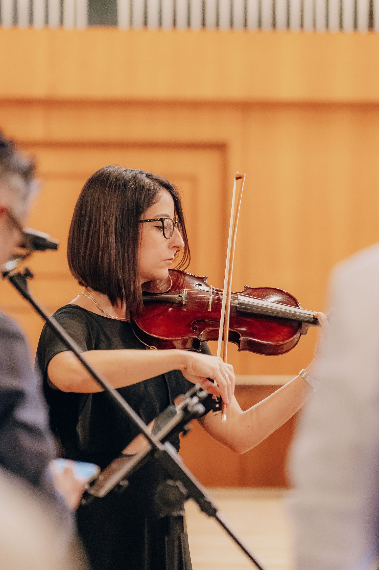 Violinist in black attire performs on stage with focused concentration in a warm-lit concert hall setting.