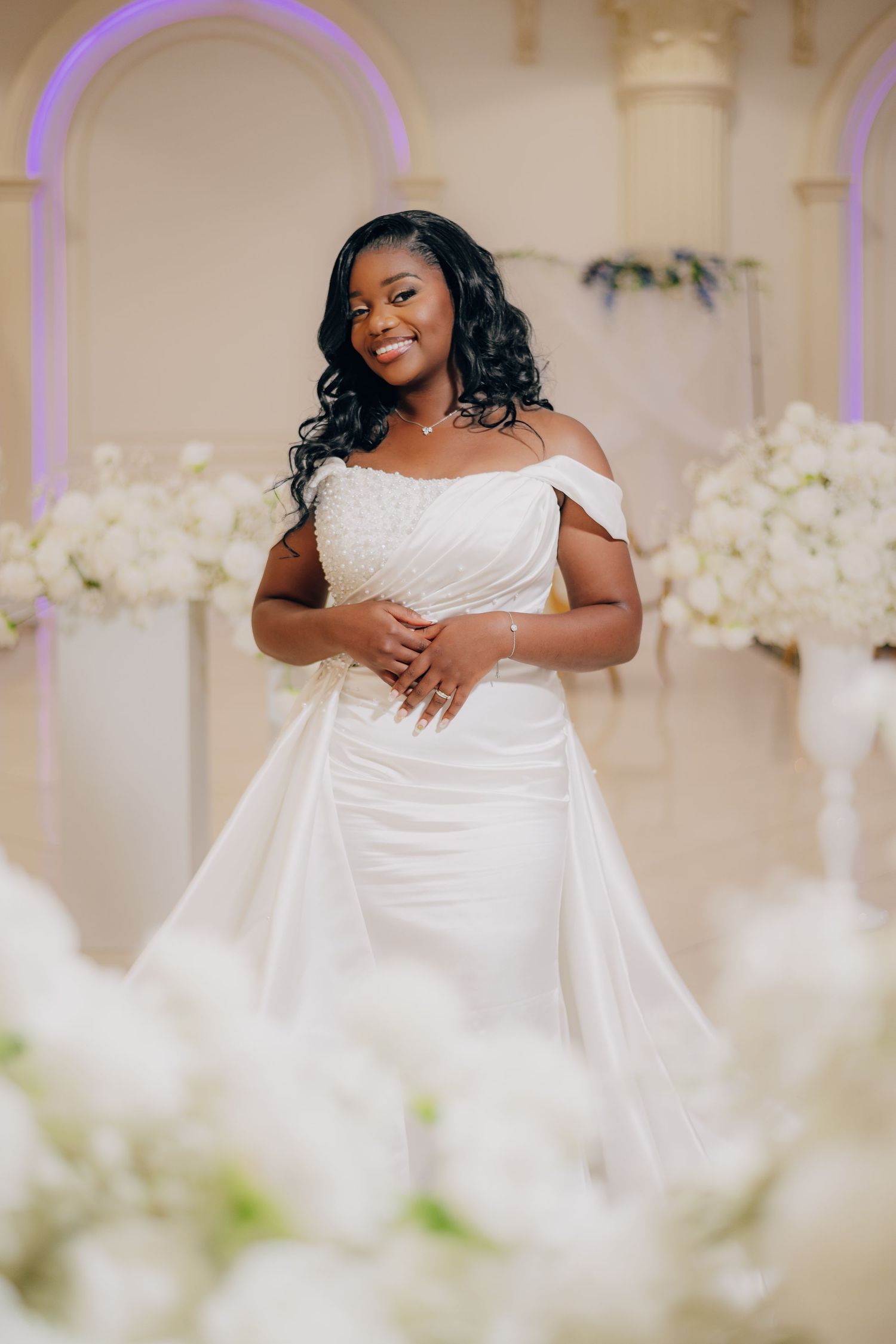 Beautiful bride in off-shoulder white wedding gown poses elegantly among white floral decorations in an ornate venue.