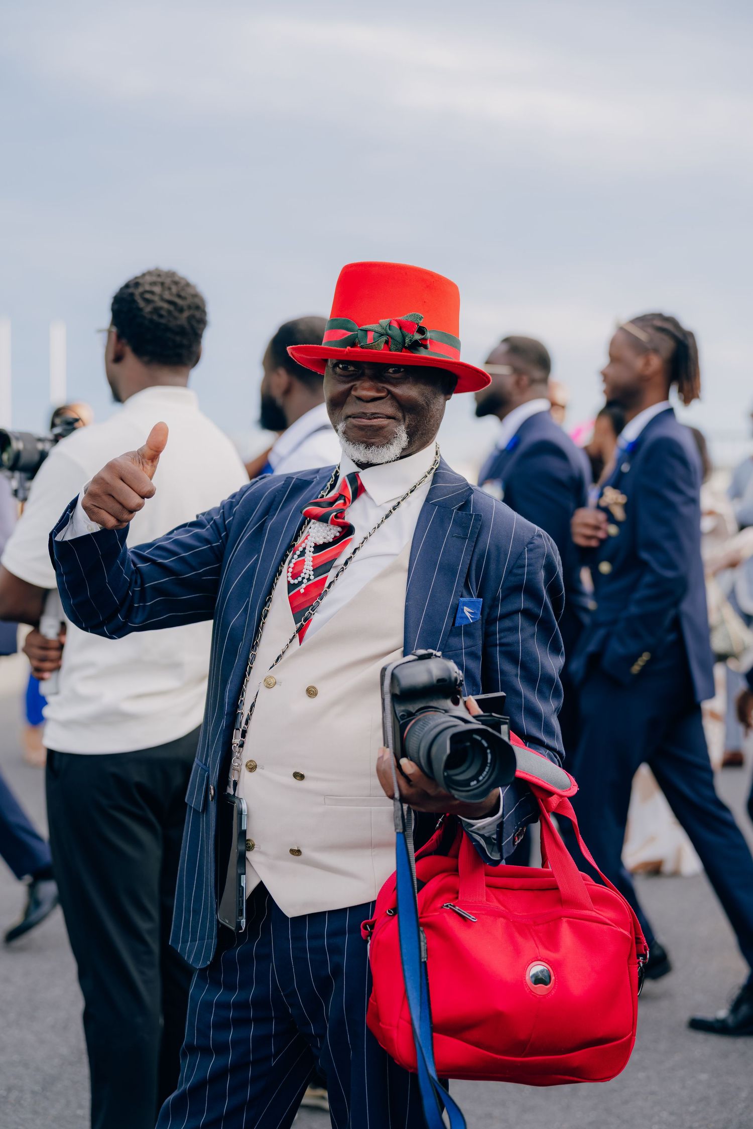 Stylishly dressed wedding guests in pinstripe suits and red accessories celebrate at an outdoor reception.