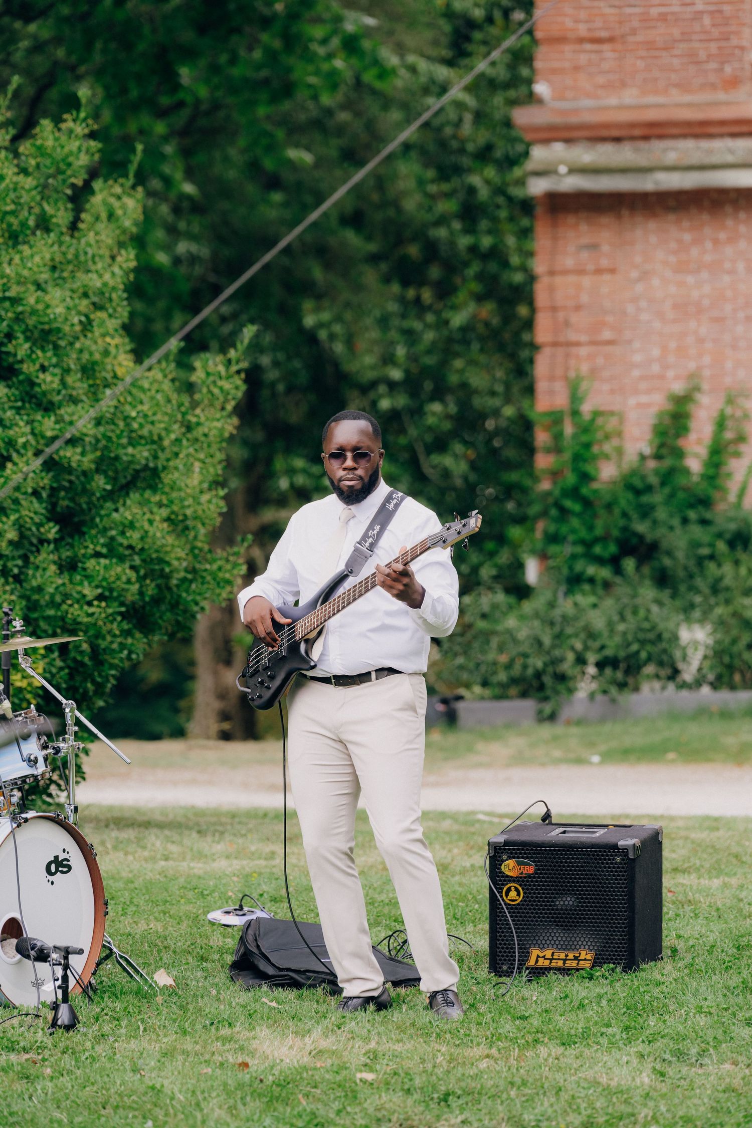 Musician in white attire plays guitar on a grassy lawn with amplifiers and equipment visible.
