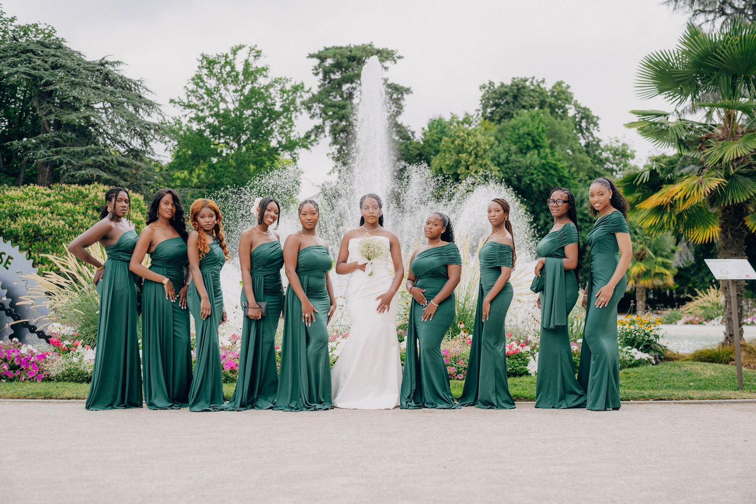 A wedding party in emerald green dresses poses together on a lush garden lawn during an outdoor celebration.