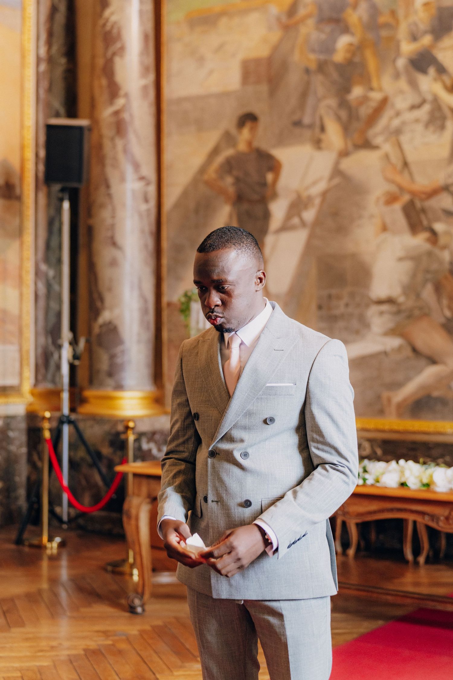 A person in a light gray double-breasted suit stands in an ornate gallery with historic murals.