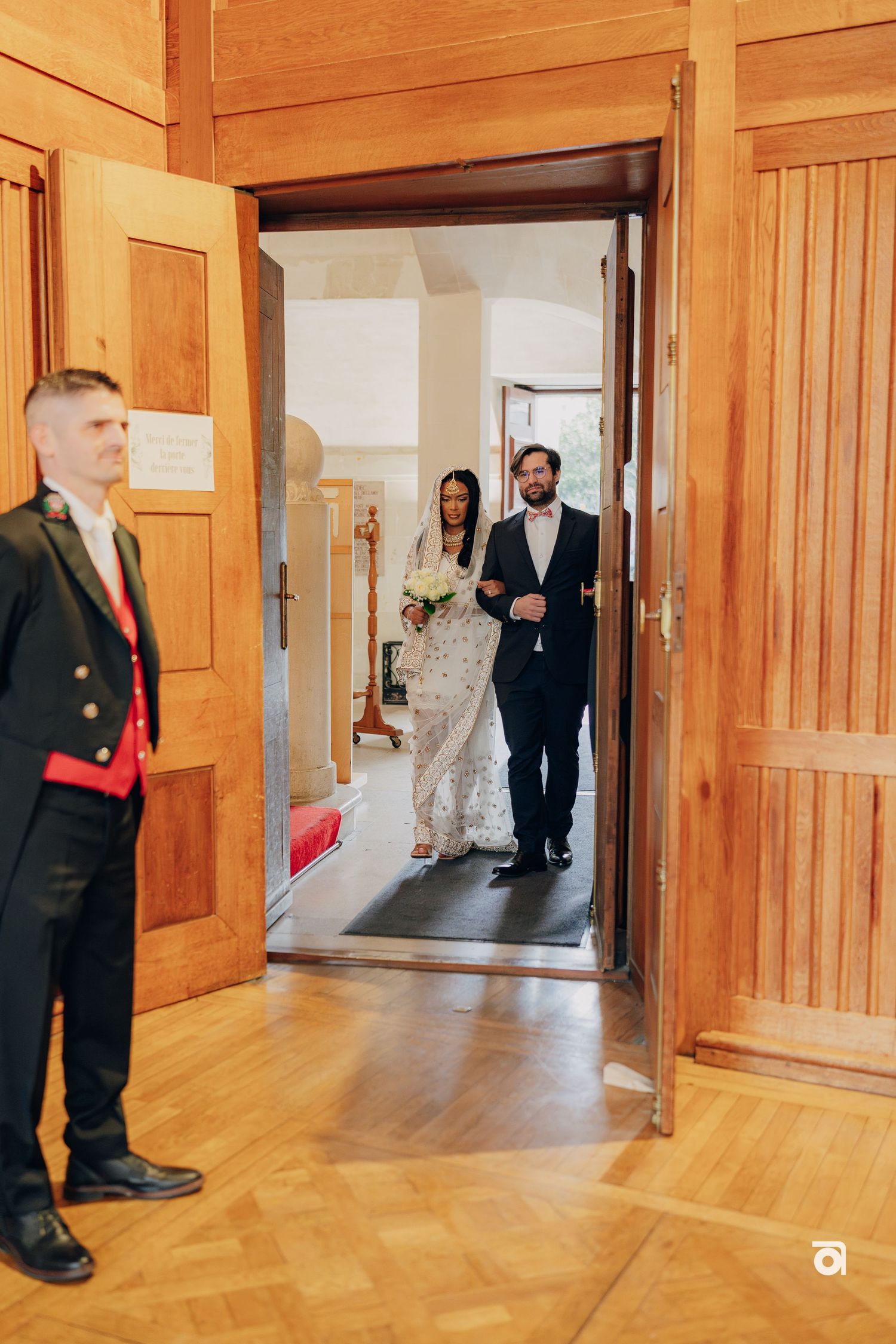 Wedding party entering through wooden doors in an elegant hallway with parquet flooring.