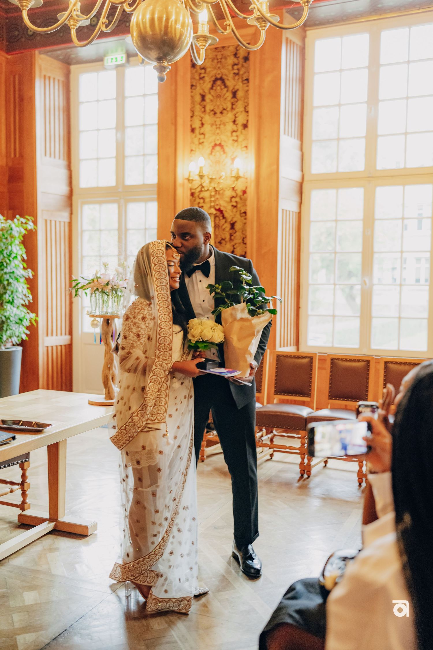 Couple celebrates during wedding reception in sunlit room with gold decor and ornate chandelier.