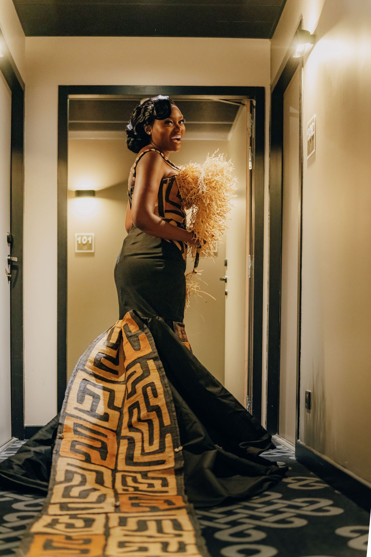 A person wearing a dramatic black dress with geometric pattern train walks down a hallway in warm lighting.