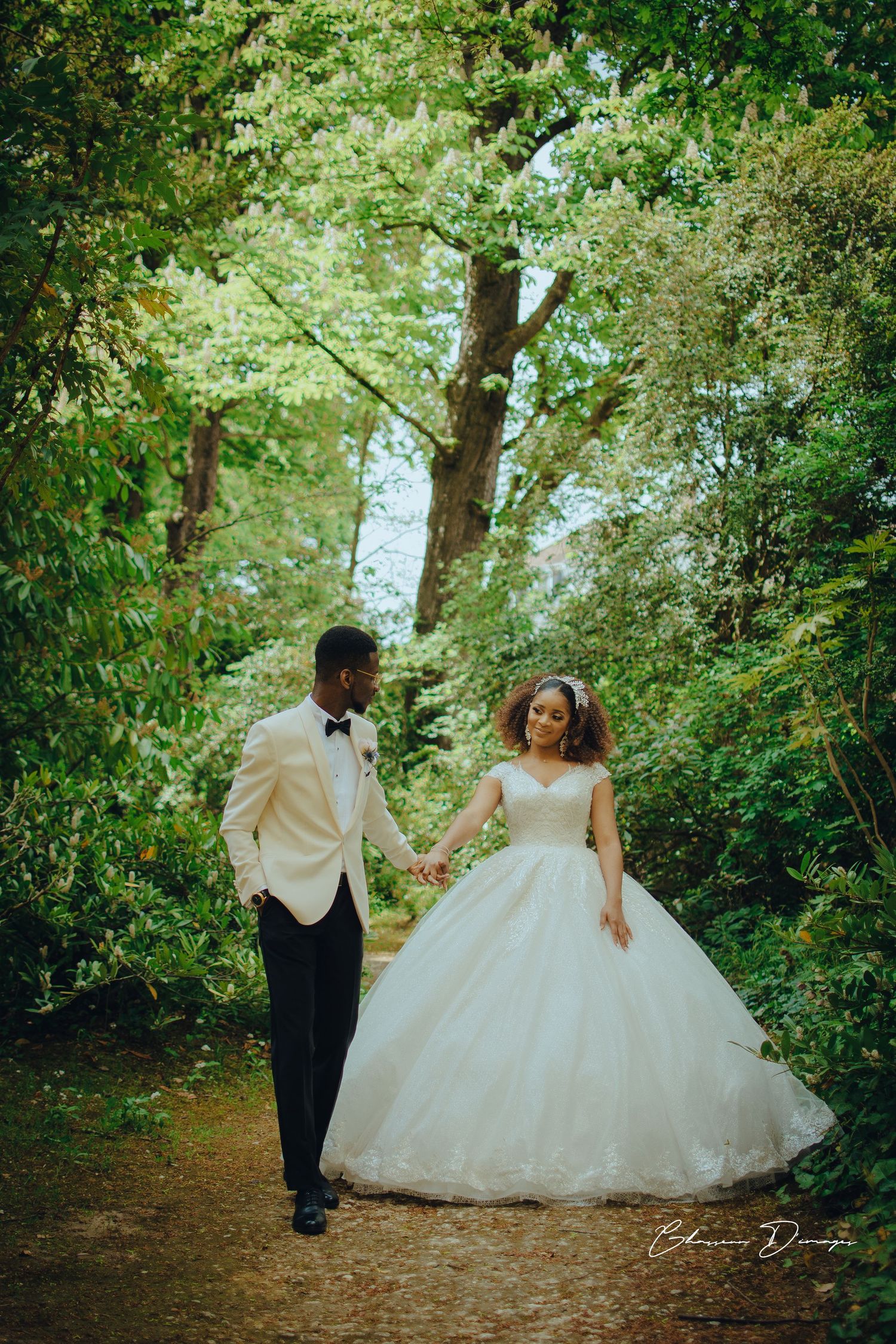 Une mariée en robe de bal blanche et un marié en smoking marchent main dans la main le long d'un sentier forestier