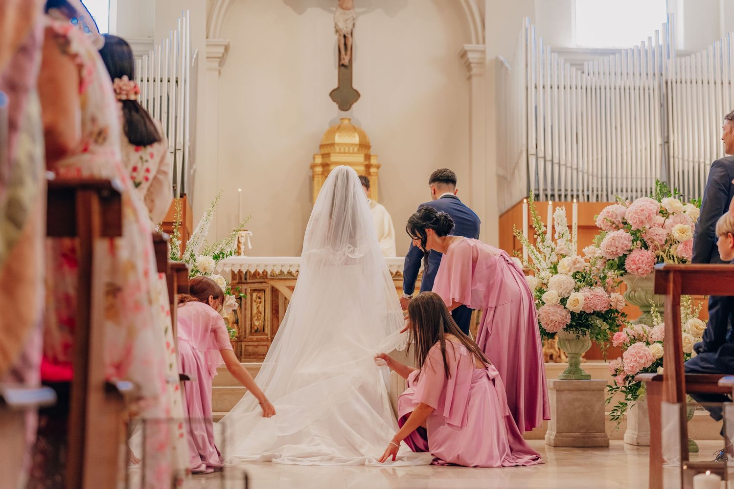 Wedding party members in mauve dresses gather around a bride in an elegant church setting with floral decorations.