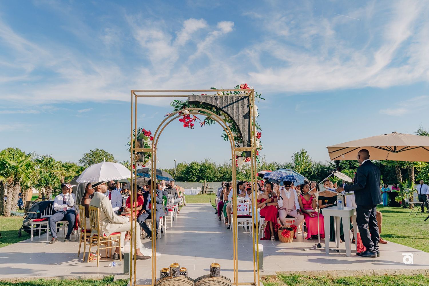 Wedding arch decorated with flowers stands against blue sky at an outdoor venue with guests seated.