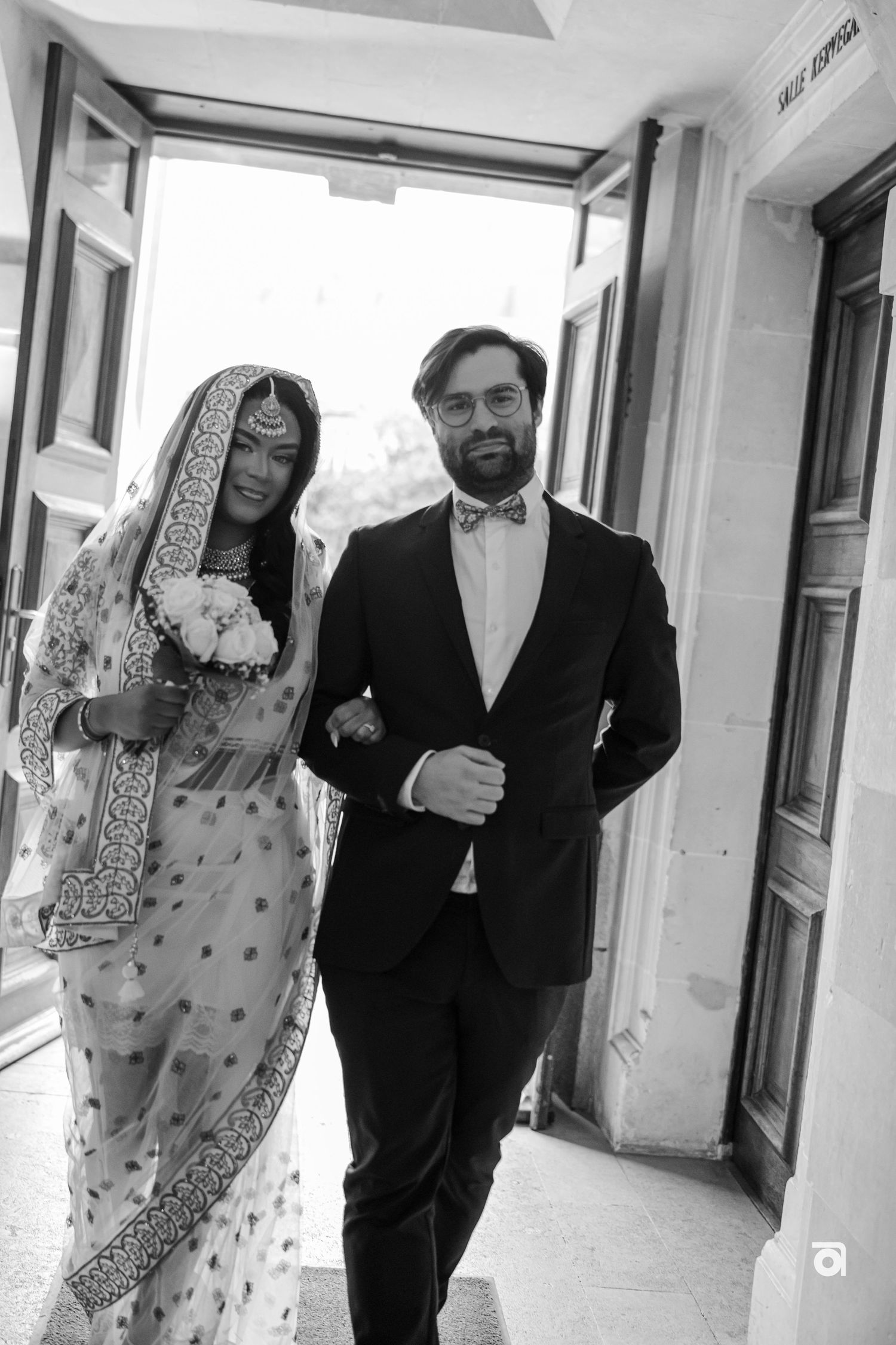 A wedding couple in traditional attire walking together, with the bride wearing an ornate sari and veil.