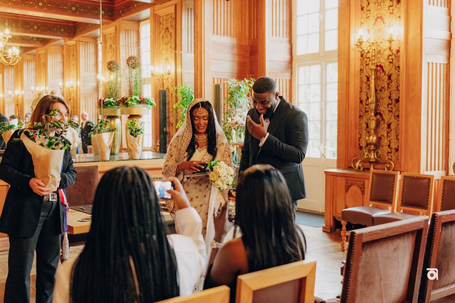 Wedding party gathers in an elegant room with gold-toned walls and natural lighting.