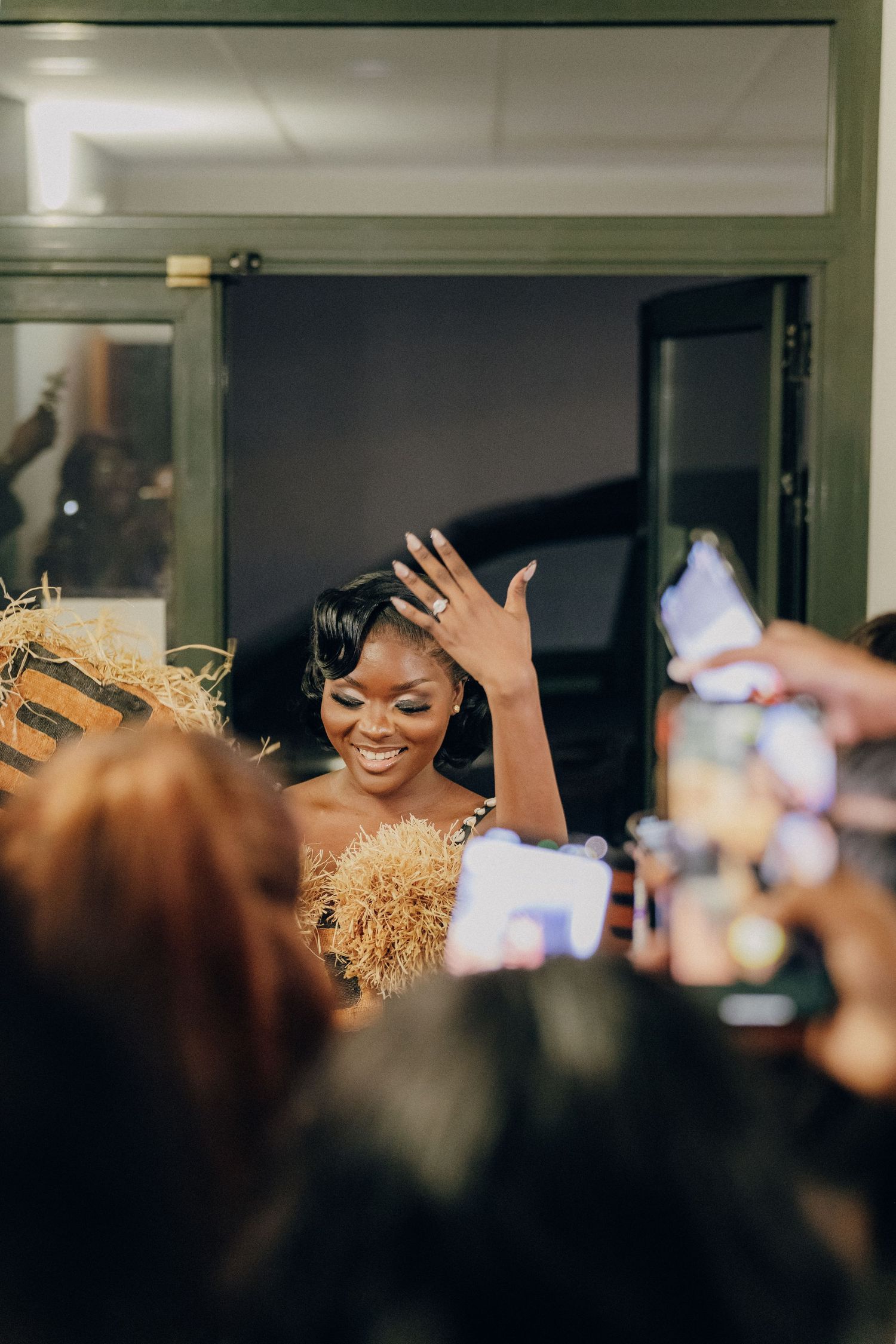 Someone in a sparkling gold outfit smiles and waves while surrounded by people taking photos on phones.