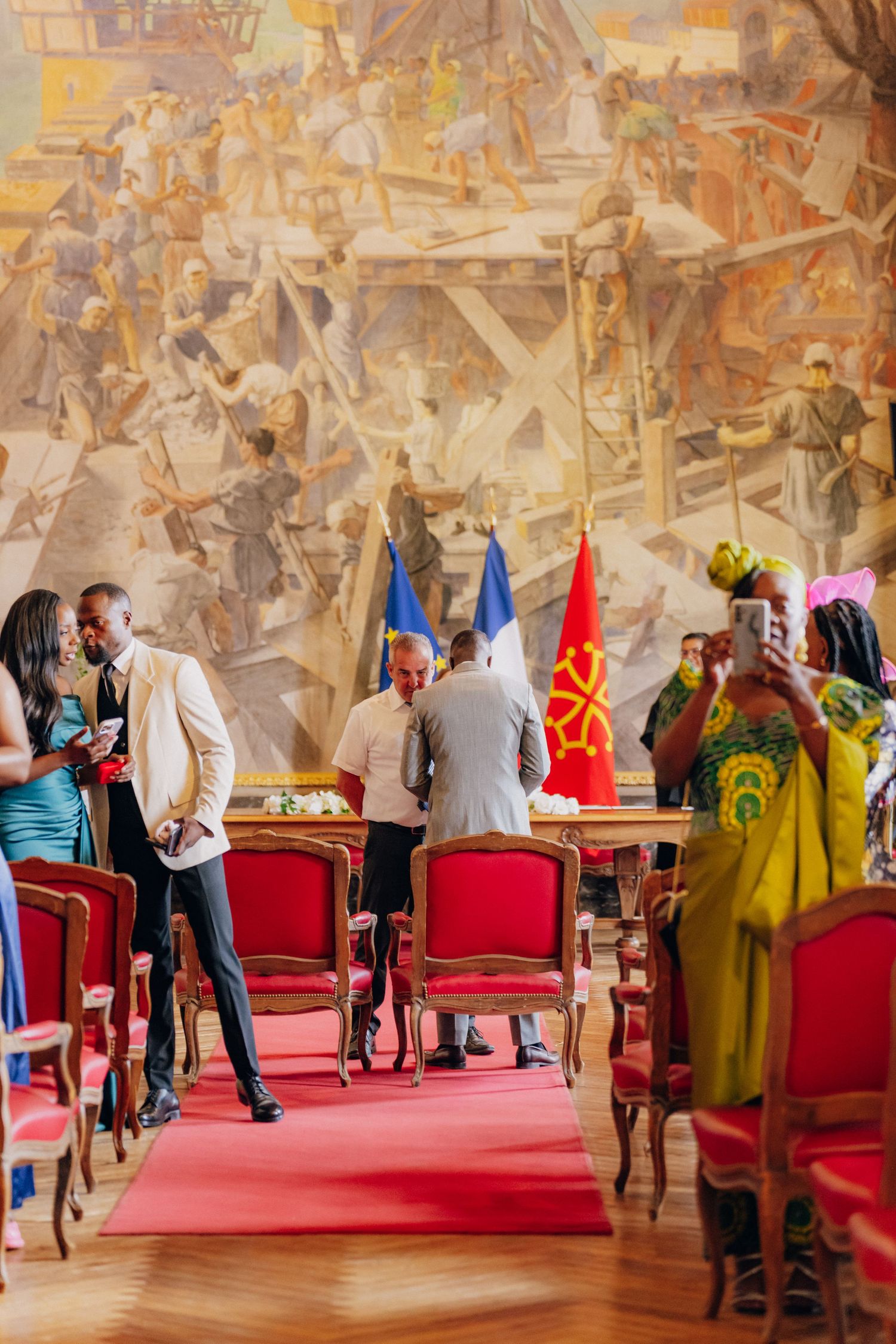 Elegant reception hall with red carpet and ornate wall murals features guests mingling around antique chairs.