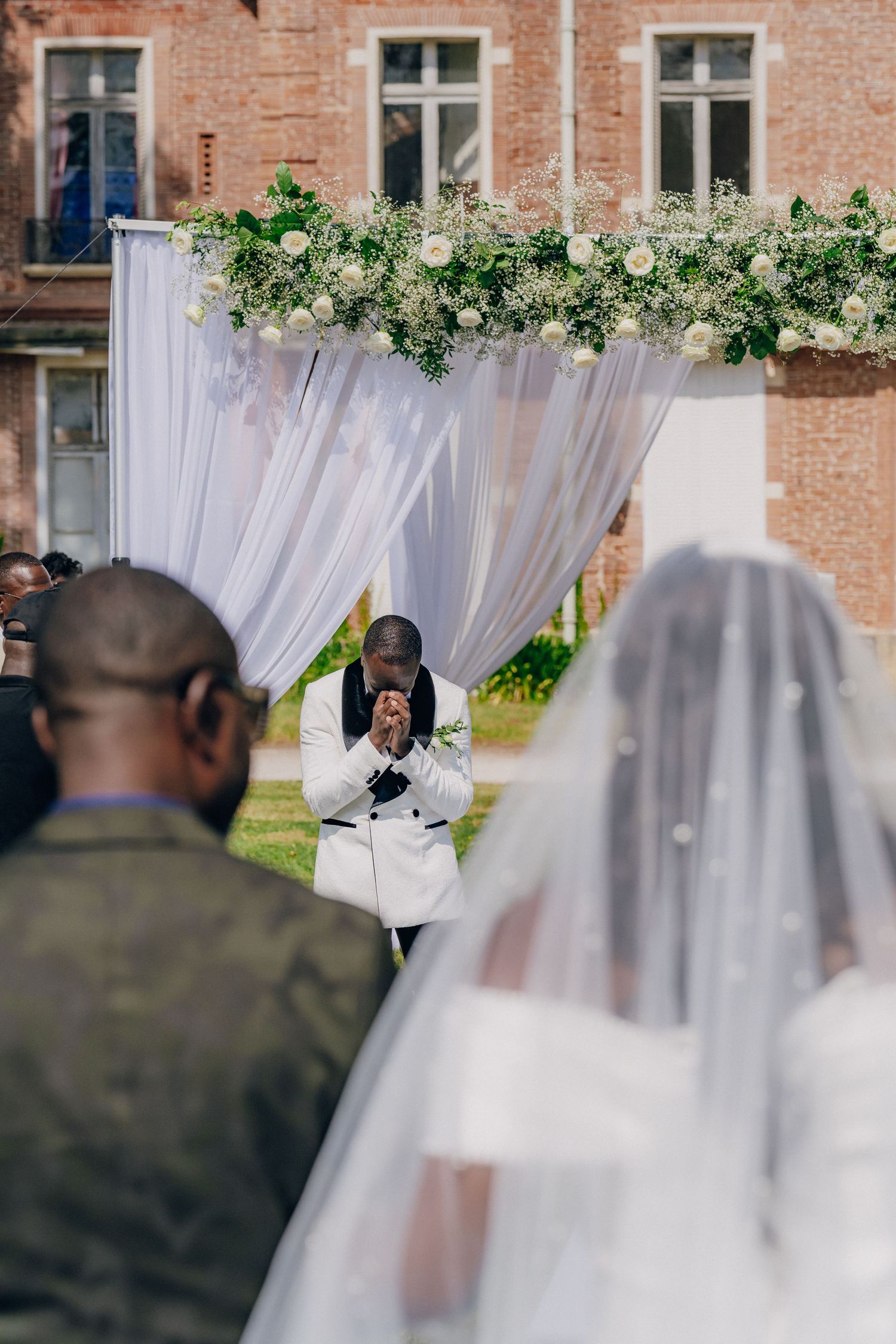Elegant outdoor wedding ceremony setup with white curtains and greenery against a brick building backdrop.