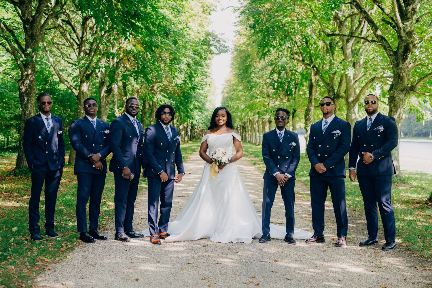Wedding party lined up on a scenic tree-lined path with groomsmen in navy and bridesmaids in neutral dresses.