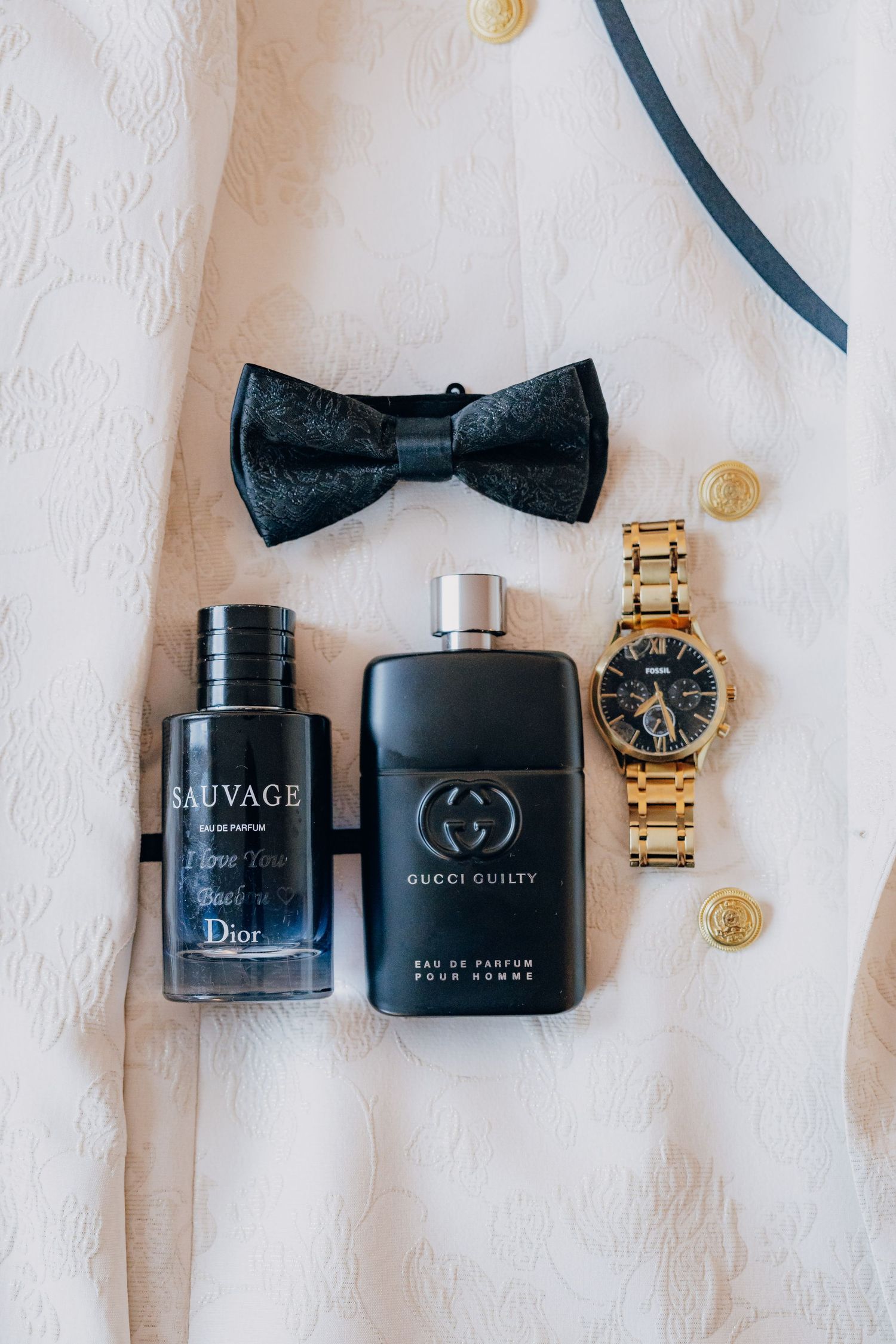 Luxury men's accessories including black bow tie, gold watch, and designer fragrances arranged on white textured surface.