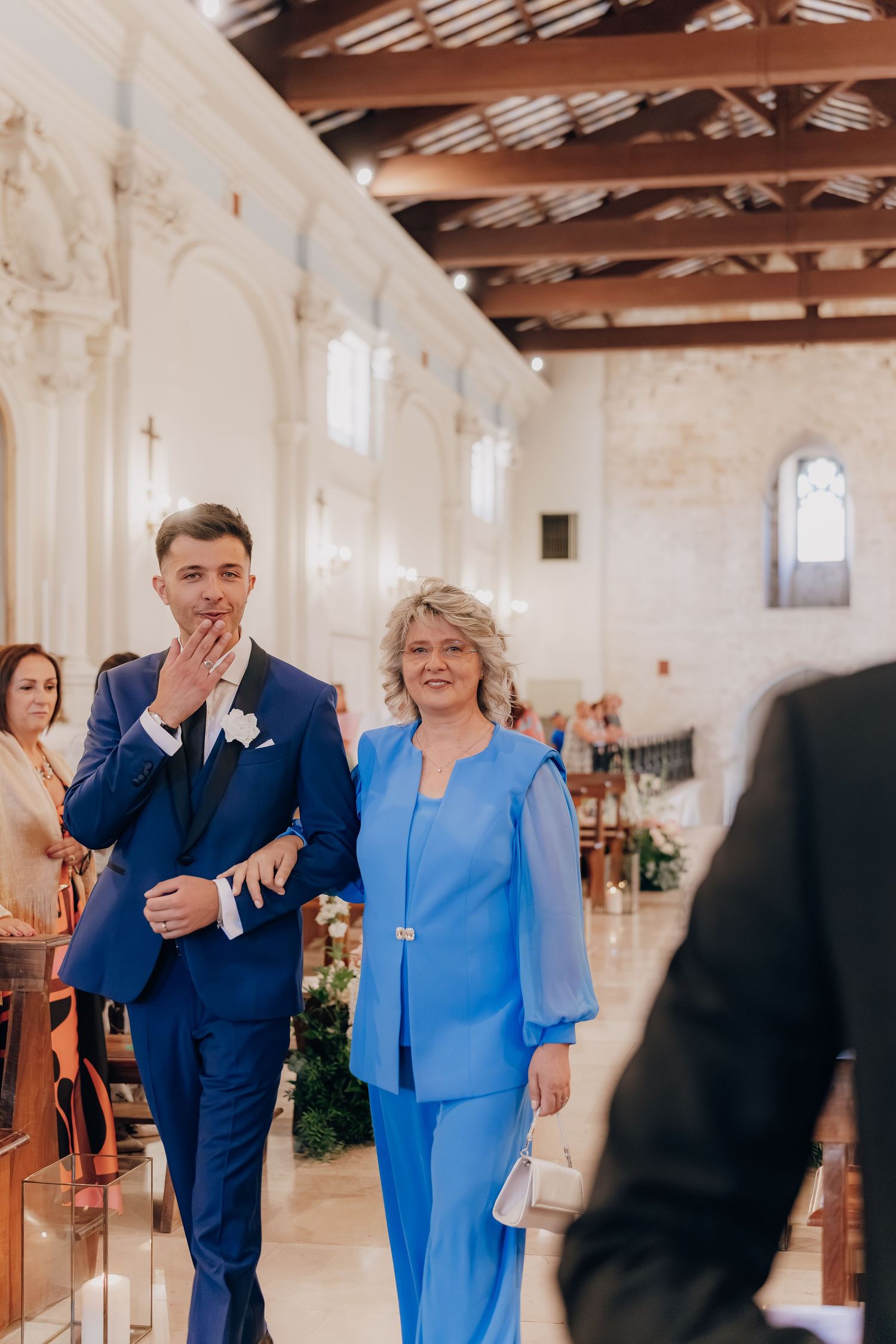Wedding party members walk through a rustic barn venue with exposed wooden beams and white stone walls.