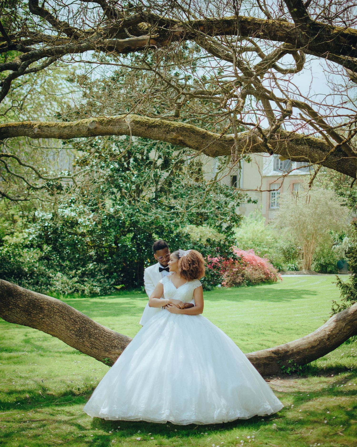 Two people in wedding attire embrace under a curved tree branch in a lush garden setting during springtime.