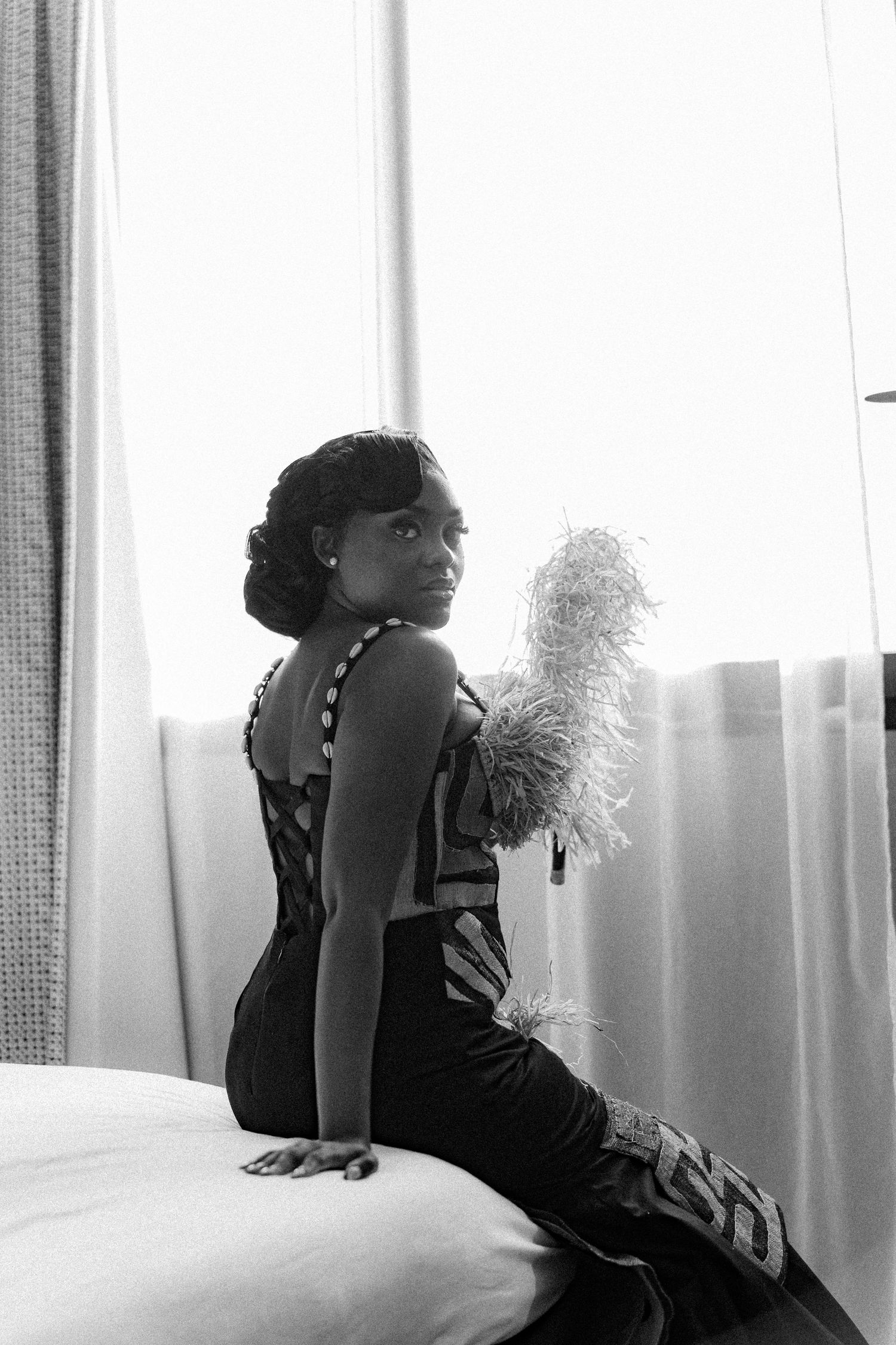 Black and white vintage-style boudoir photograph showing a figure in evening attire posing elegantly on furniture by a curtained window.