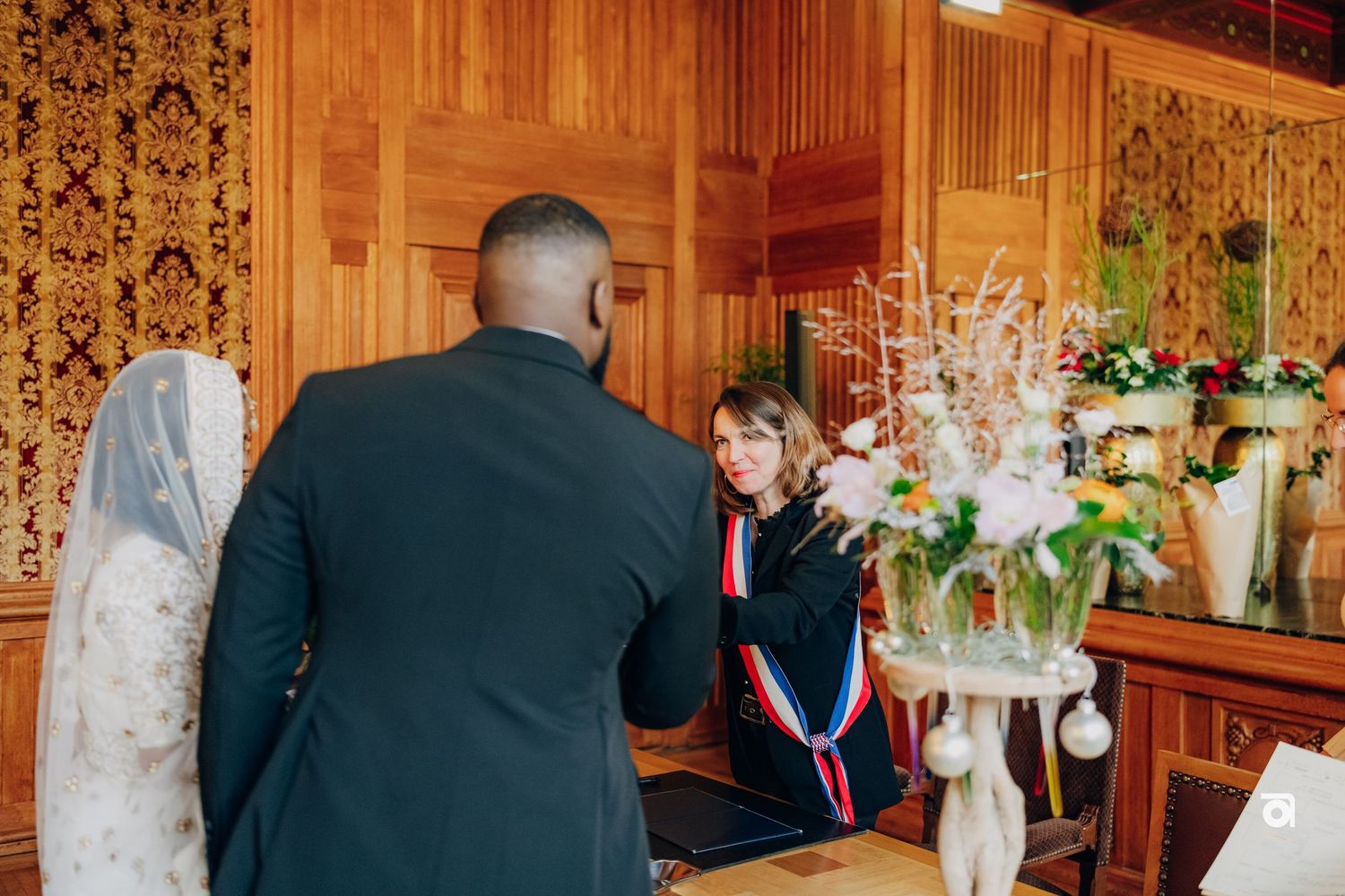 A wedding ceremony in a wood-paneled room with floral arrangements and official in traditional dress.