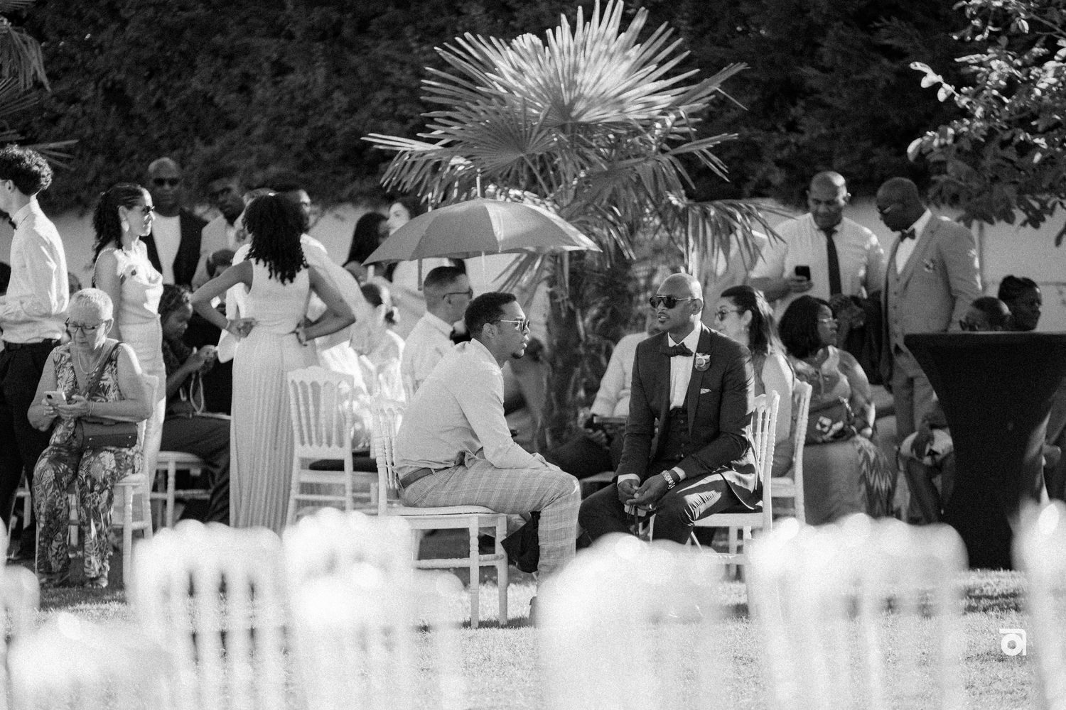 A crowd gathers for an outdoor garden party with umbrellas and white chairs visible in black and white photography.