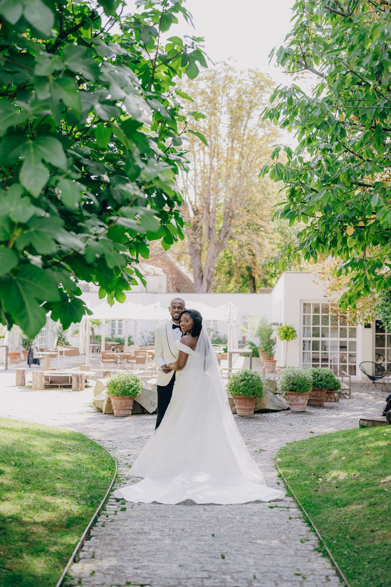 A newlywed couple strolling down a stone pathway lined with trees and manicured gardens.