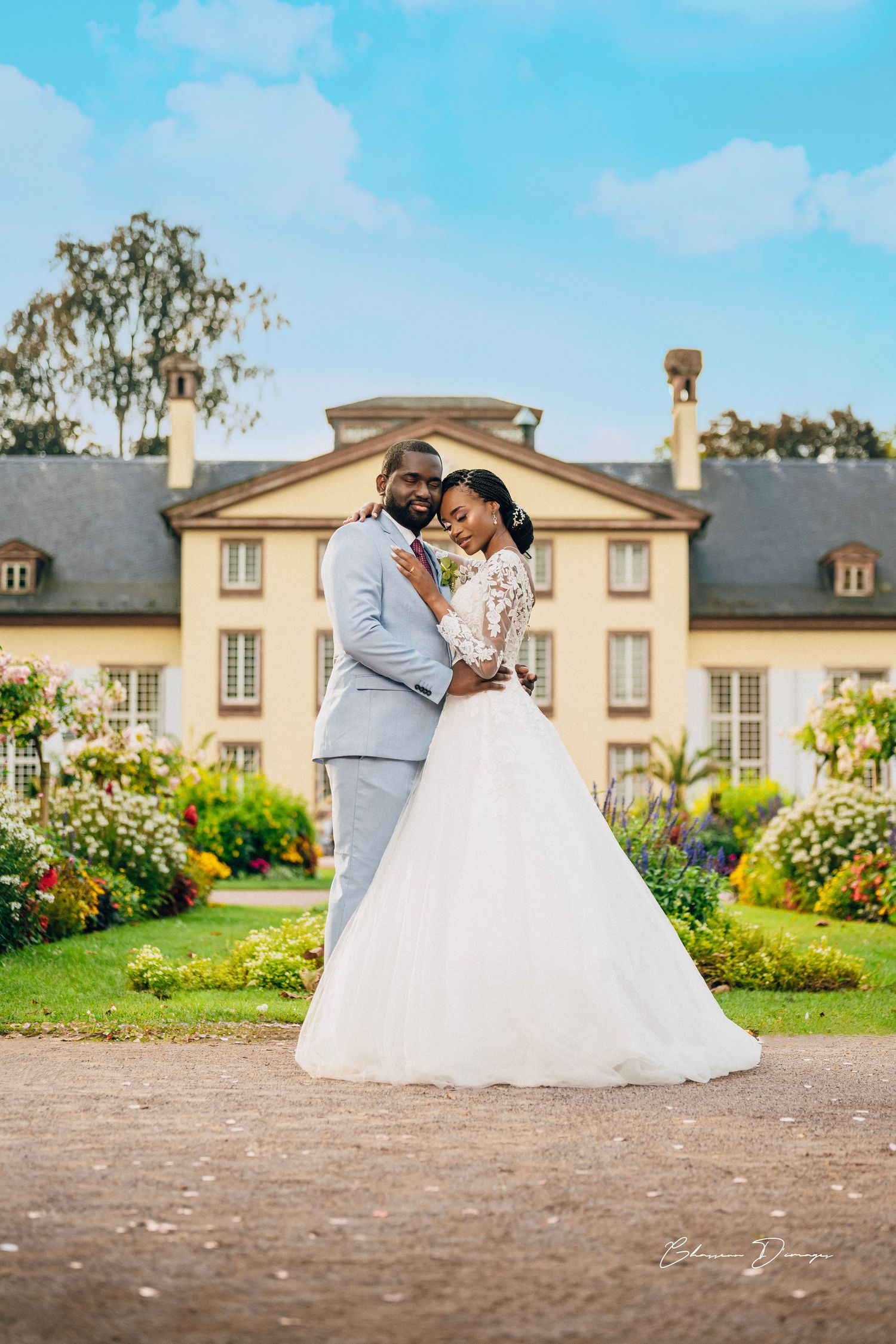 A happy couple embraces in their wedding attire in front of a grand estate with beautiful flower gardens.