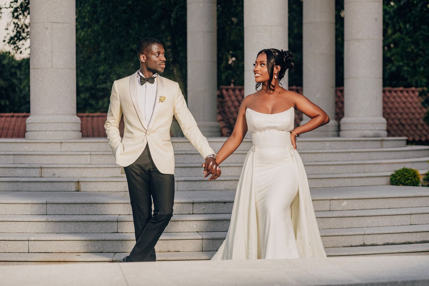 Couple in formal attire holding hands on marble steps with classical columns.