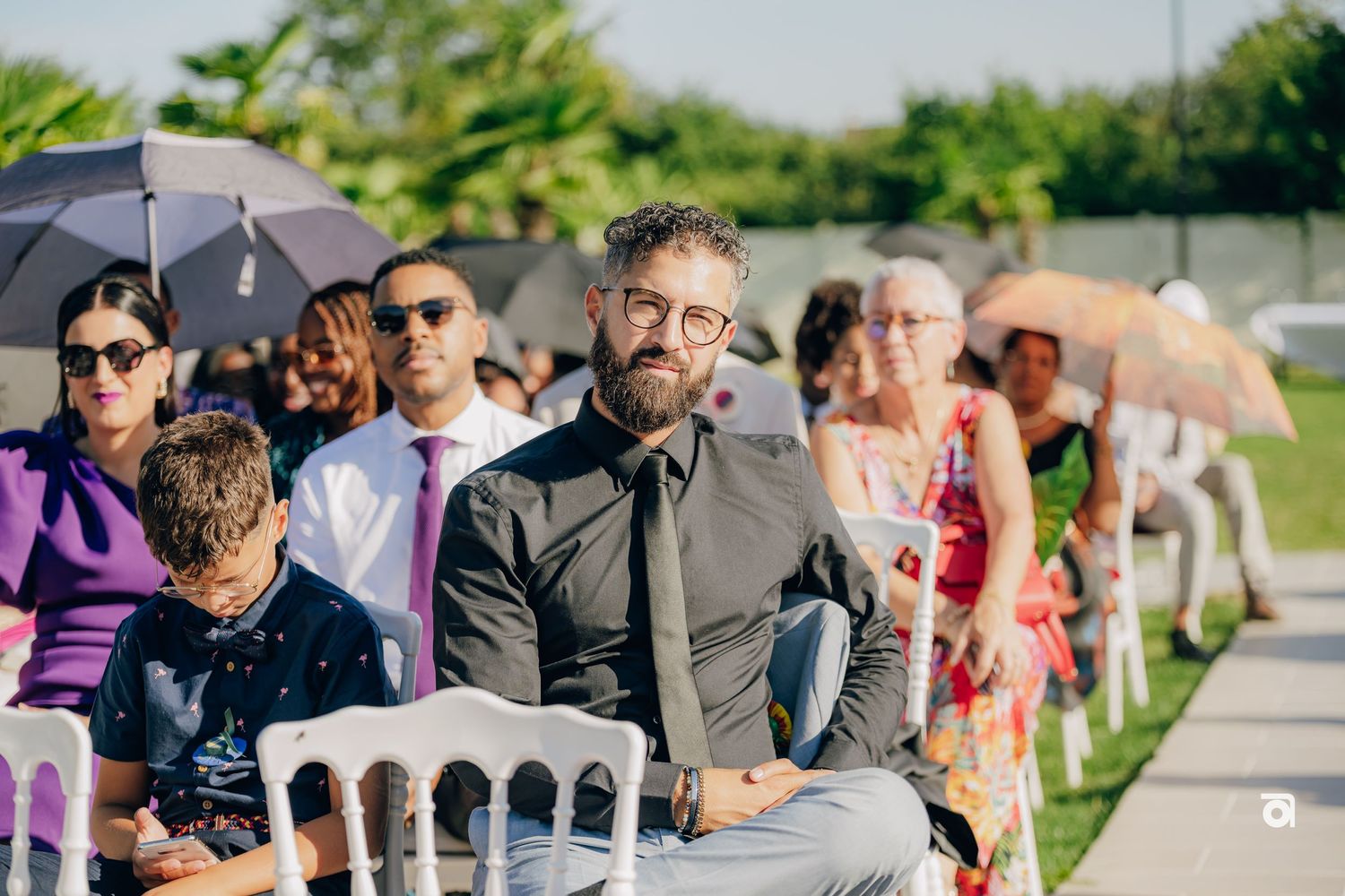 Outdoor wedding ceremony with guests seated in white chairs under umbrellas.