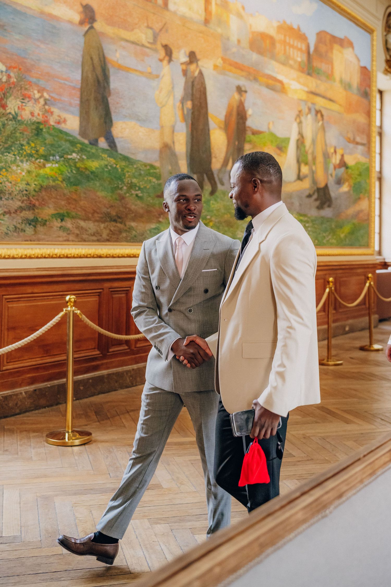 Two well-dressed people in light suits converse in an elegant gallery with classical murals on the wall.